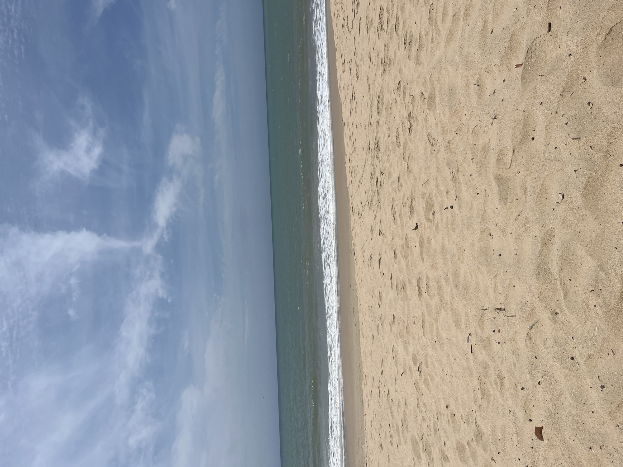 The image depicts a beach scene in La Pared, Venezuela. The view is oriented vertically, showing a section of sandy beach on the right, the ocean in the middle, and a blue sky with scattered clouds on the left. The sand is light tan and marked with footprints and small debris. The ocean is a greenish-blue, with white foam from the waves meeting the shore. The sky shows a gradient of blue, with wispy, white clouds spread across the upper portion. No people are visible in the image.