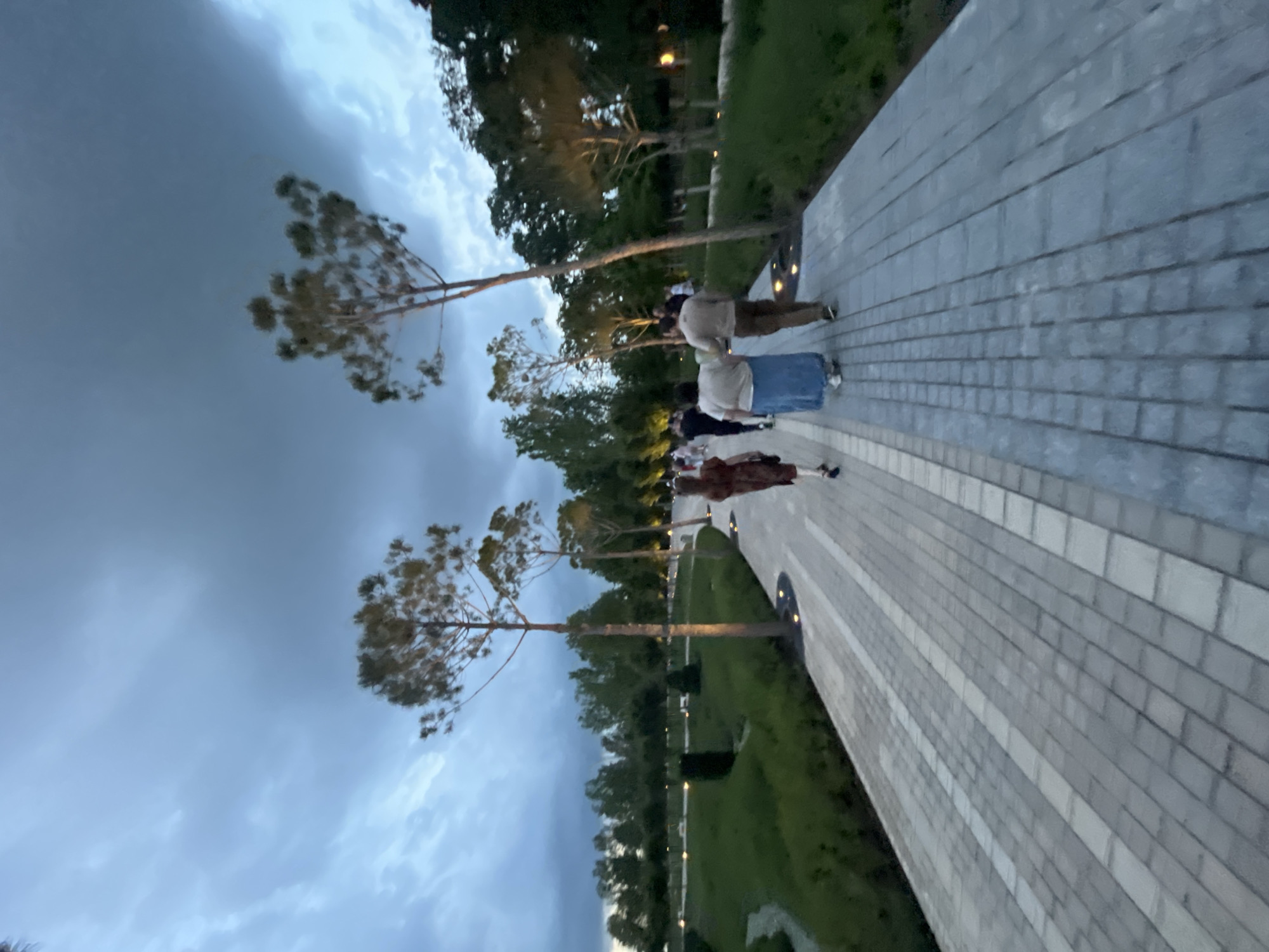 The image shows a paved walkway with a mosaic pattern of light and dark gray bricks, lined with grass and trees on either side. Several people are walking away from the camera on the walkway. The sky is dark and cloudy, suggesting it might be evening or a storm is approaching. In the background, there are more trees and some subtle lighting. The scene appears to be a park or a landscaped outdoor area. Based on the visual information, this could be a scene from the Park of Eternal Flame (Park Pobedy) in Krasnodar, Russia, which features similar paved walkways and is a popular public space.