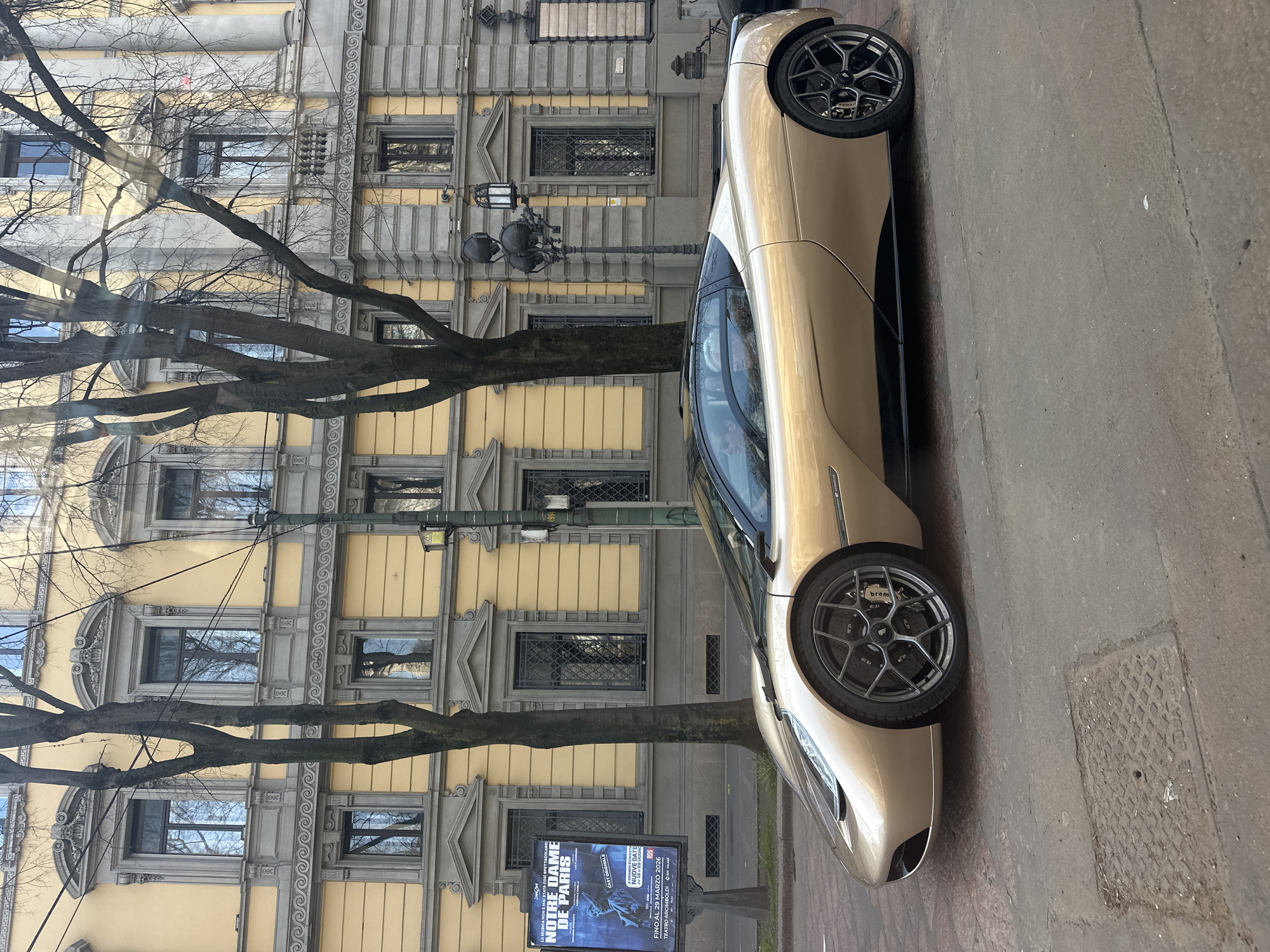 A sleek, gold-colored two-door sports car with a black roof and dark multi-spoke wheels, featuring visible 'Brembo' brake calipers, is parked on a cobblestone-like paved street in Milan, Italy. Two occupants are visible within the vehicle's tinted interior. The background features a multi-story building with a light yellow facade extensively detailed with grey classical architectural elements, including cornices, ornate window surrounds, and balustrades. Several bare deciduous trees with dark trunks line the street, partially obscuring the building. To the left of the car, a large blue billboard advertises "Notre Dame de Paris" at "Teatro Arcimboldi - Milano," with dates indicating performances until March 29, 2026. An ornate black street lamp pole with multiple lanterns stands on the right. A metal manhole cover is present on the pavement in the foreground.