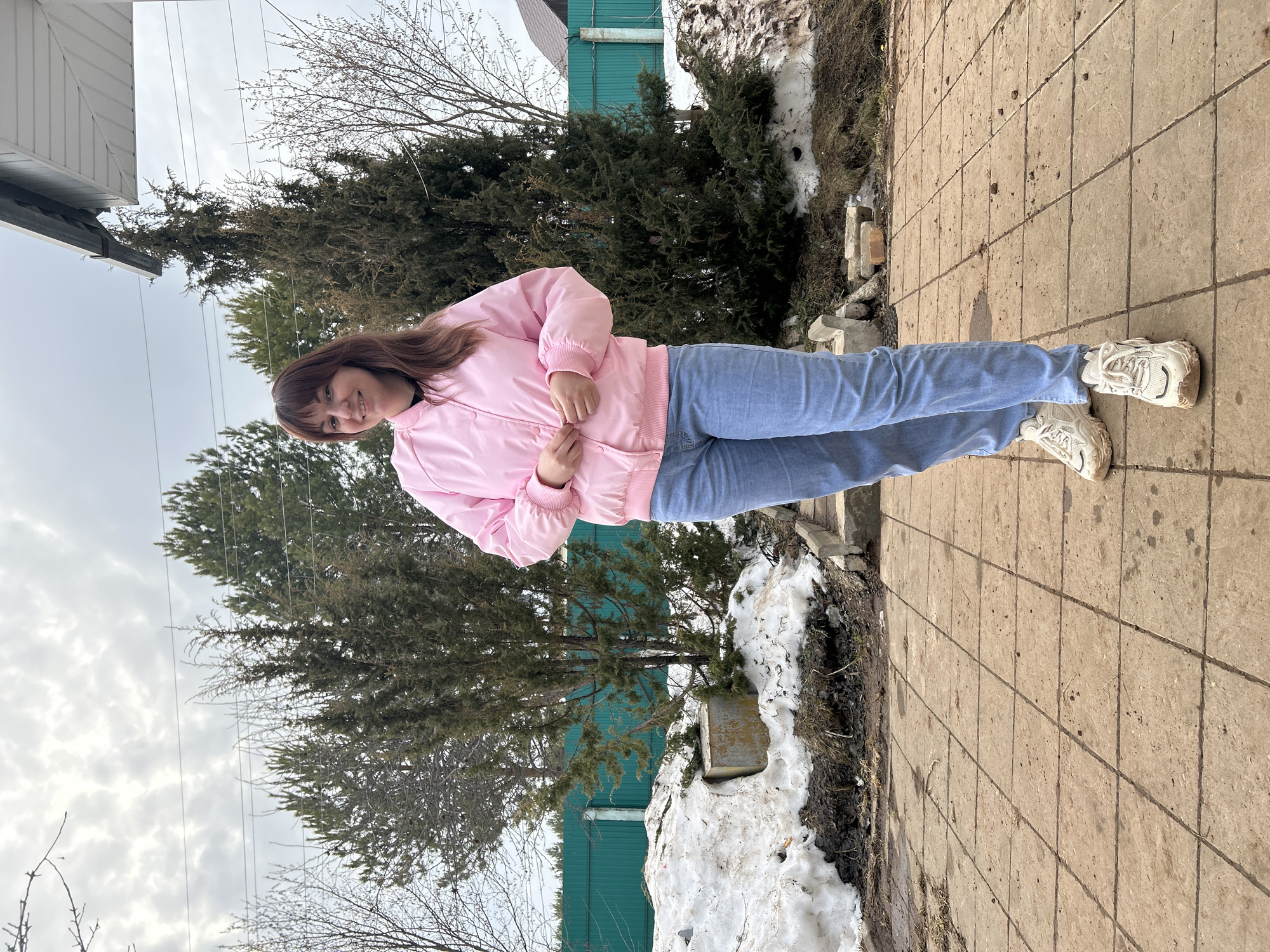 A full-body photograph depicts a smiling Caucasian female subject outdoors in Фоки, Russia. The woman has medium-length brown hair with bangs and is positioned centrally, facing the camera. She wears a light pink bomber jacket, a dark shirt underneath, light blue jeans, and chunky white sneakers showing dirt. Her hands are clasped together at waist level, appearing to adjust the jacket. She stands on a light brown paved surface composed of square tiles, which exhibits scattered dirt and debris.
To the left of the subject, a patch of melting snow is present, mixed with dark earth and sparse brown vegetation. A partially visible, rectangular concrete or metal box is embedded in the snow. In the mid-ground, a dark green corrugated metal fence extends horizontally. Numerous conifer trees, varying in shade from dark green to brownish-green, are visible behind the fence and to the subject's right. Bare deciduous tree branches are interspersed throughout the scene. An overhead white building soffit and gutter system is visible in the upper right quadrant of the image. The sky is overcast, and power lines traverse the upper portion of the frame. The environment indicates a transitional season, possibly late winter or early spring, given the presence of snow and bare trees.