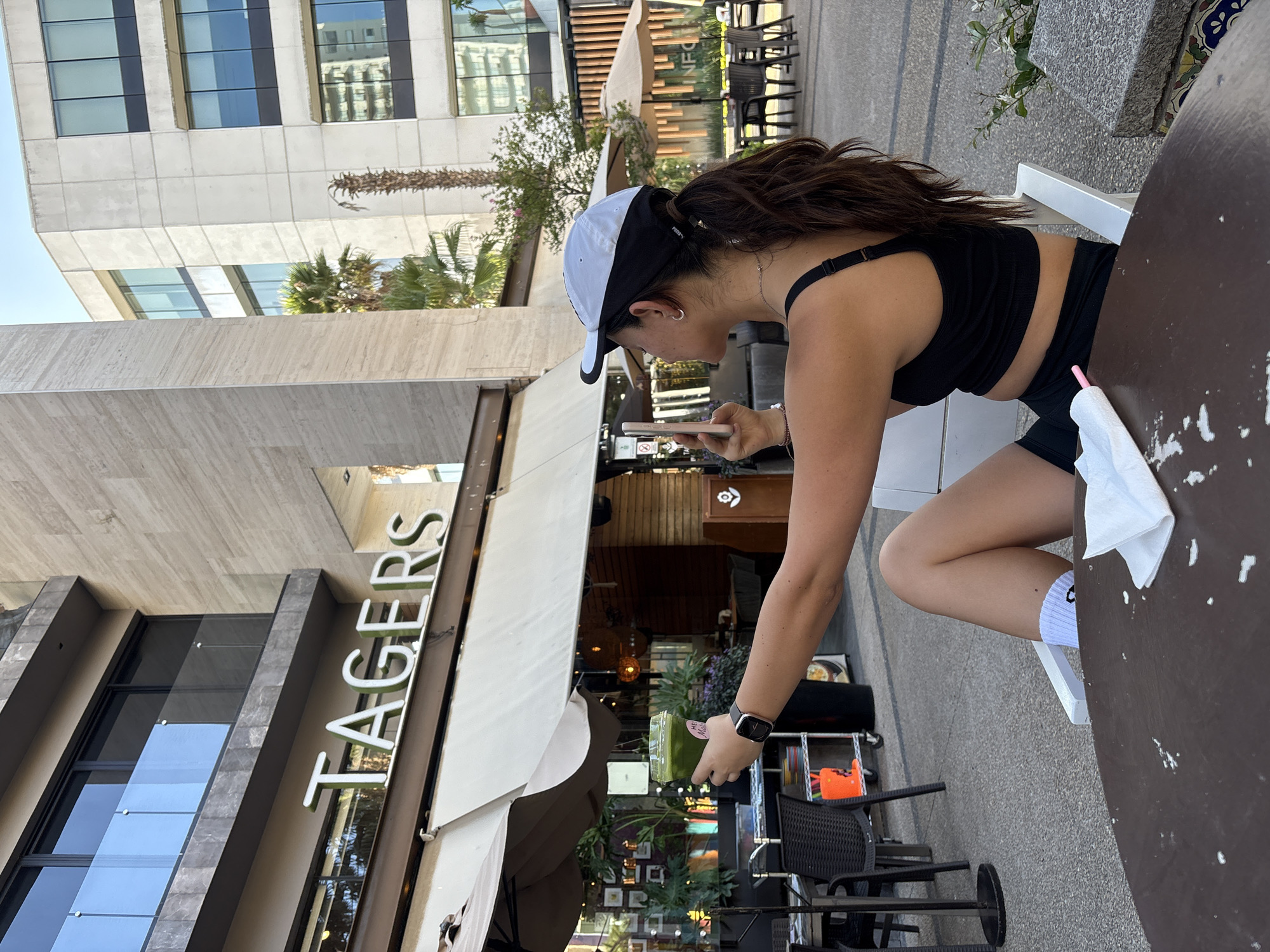 A medium shot depicts a woman at a cafe in what appears to be an urban environment. The cafe's name, "TAGERS," is visible above an awning. The woman, dressed in a black athletic top and shorts, sits at a round table on a white chair. She is wearing a black and white baseball cap and has dark hair pulled back in a ponytail. She is holding a cell phone in one hand and a drink in the other. Her arm holding the drink extends towards a table. The table has a white napkin on it. The background includes part of a building with glass windows, palm trees, and outdoor seating.
The location is San Bernardino Tlaxcalancingo, Mexico.