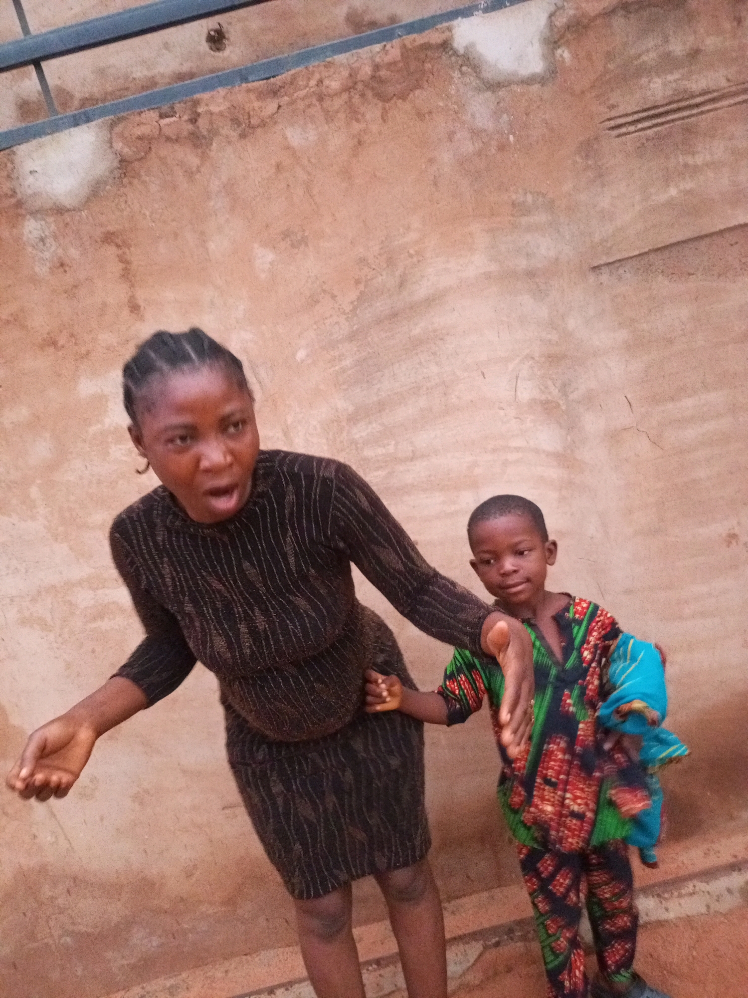 A medium shot shows a Nigerian woman and a young boy standing against a textured, light brown wall. The woman is on the left, facing forward with her mouth open, as if speaking or yelling. She has braided hair and is wearing a dark, textured dress with vertical lines. Her left arm is extended, with her hand open. The boy is on the right, looking slightly to his left, with his right arm extended, his hand open and pointing. He is wearing a colorful, patterned outfit with green, red, and black designs, and a blue cloth is draped over his left arm. The wall behind them appears to be an outdoor structure, possibly a building facade or boundary. The image is likely taken in an urban or semi-urban environment within Tempure, Nigeria.