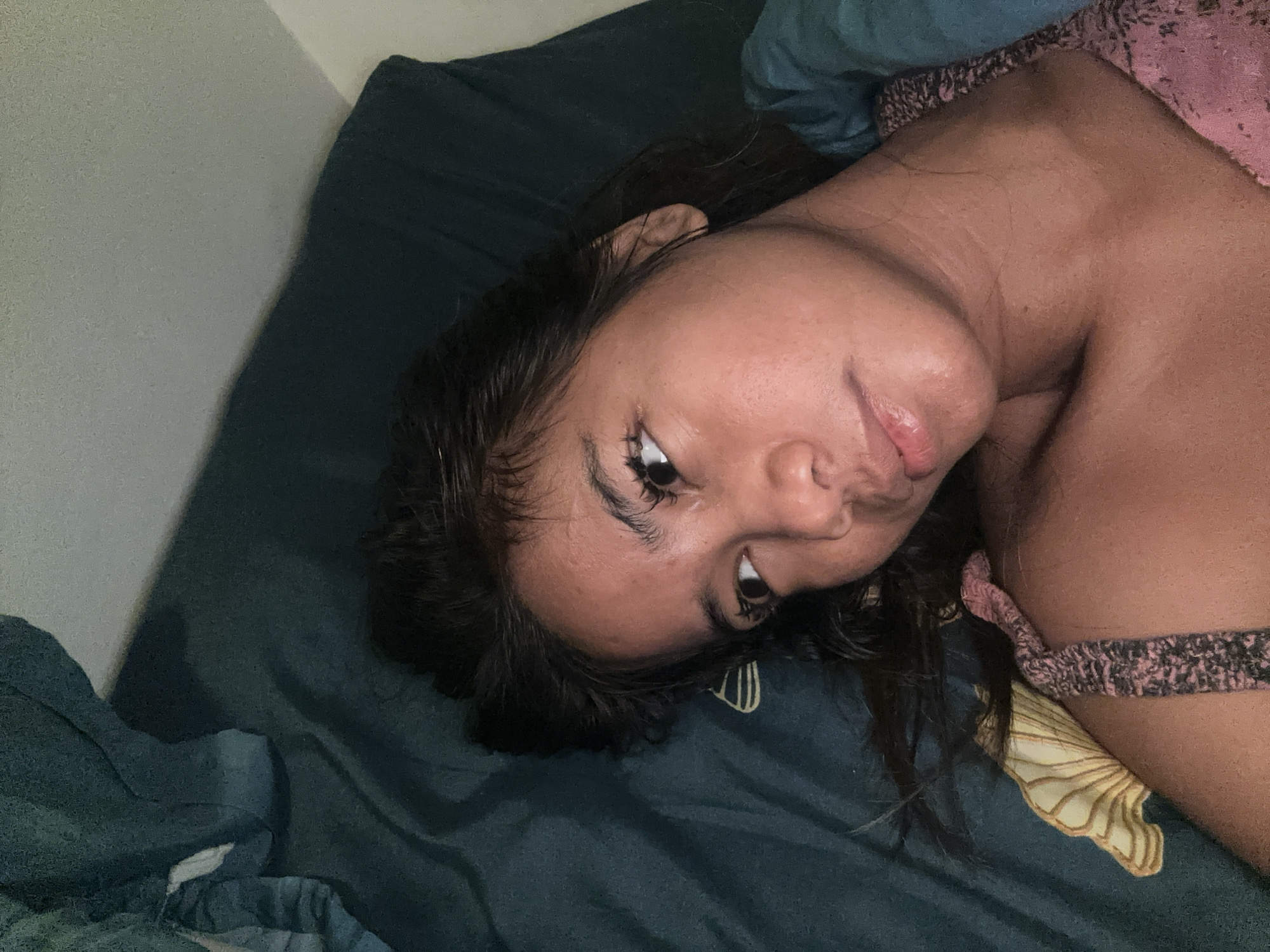 A person is lying on their side in bed, their head tilted to the side. The room is dimly lit. The person is wearing a pink garment with a patterned strap. The bed appears to be covered in a dark green comforter. The wall behind the person is a light gray.