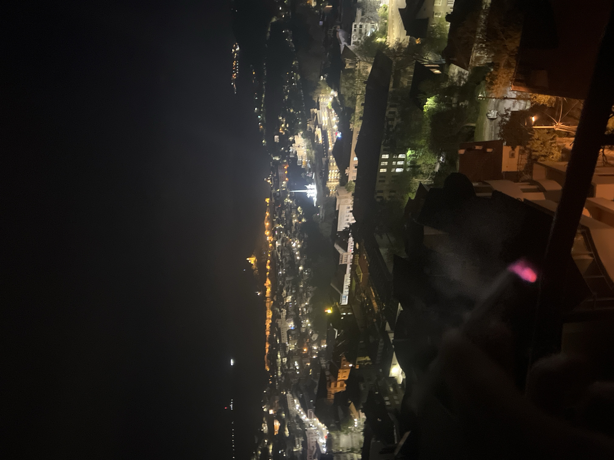 An eye-level, vertical night shot captures a nighttime aerial view of Veliko Tarnovo, Bulgaria. Illumination from artificial lights dot the landscape, creating a dense network of shimmering points amidst the darkness. These lights emanate from buildings and line the pathways, delineating the city's outline and architectural structures. The cityscape extends into the horizon, where the lights gradually fade into the blackness. In the immediate foreground, out-of-focus architectural structures rise, partially obstructing the view. A hand is also visible in the foreground, holding what appears to be a lit cigarette, with a pink ember. The overall composition suggests an elevated observation point, offering an expansive panorama of the city.