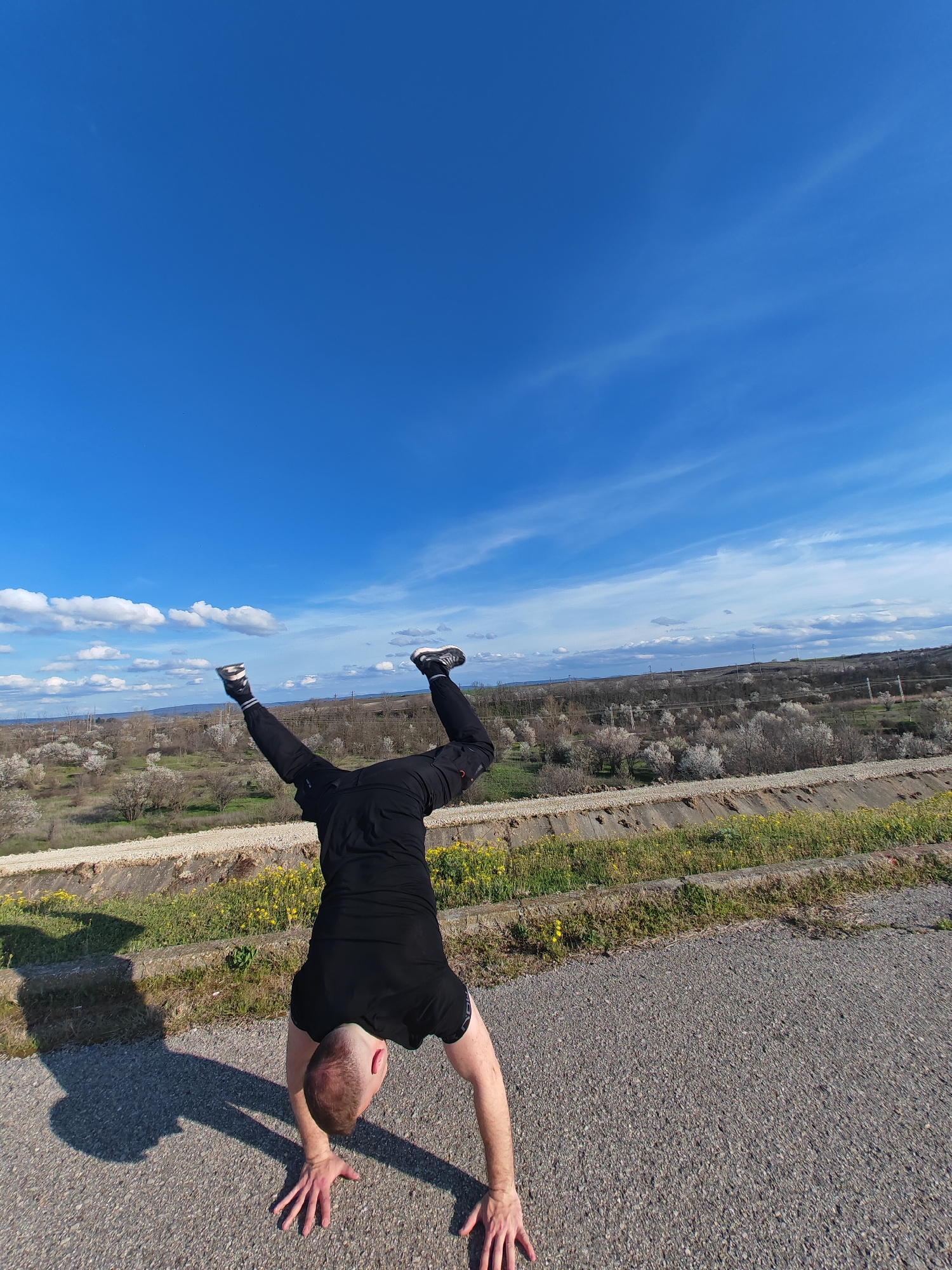 A man is performing a handstand on an asphalt road. He is wearing black athletic clothing and athletic shoes. Behind him, a vegetated embankment stretches towards a landscape of flowering trees and shrubs. The sky above is blue with scattered clouds. The location is Shumen, Bulgaria.