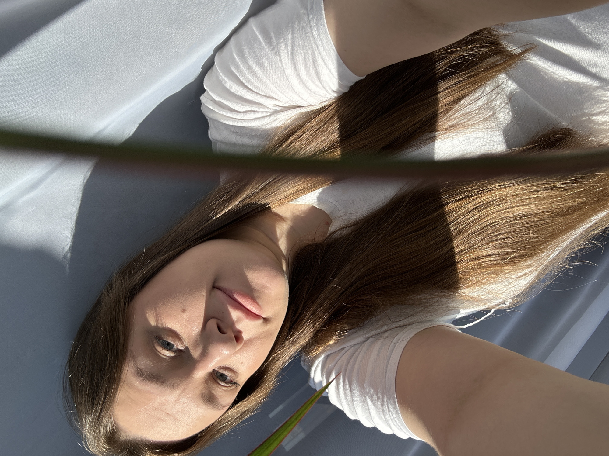 A woman with fair skin and long brown hair is seen in a selfie-style shot, appearing to lie down. She wears a white t-shirt. Sunlight creates highlights and shadows on her hair and face. A plant with a green stem and leaves is partially visible near her face. The background appears to be a light grey fabric. There is no location visible to determine a location in the city of Ольховка, Russia.