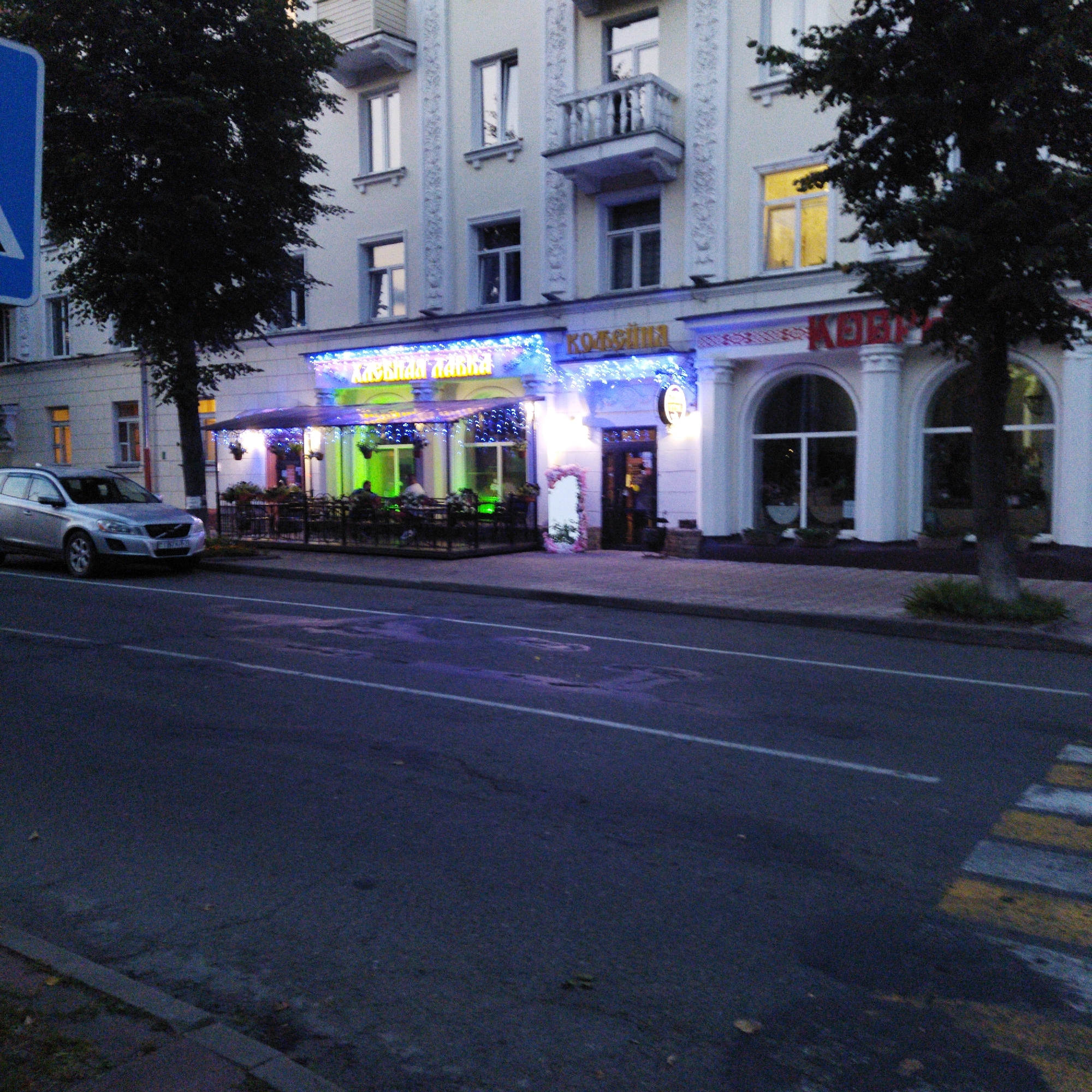 The image depicts a street scene in Polotsk, Belarus, during twilight. A silver Volvo XC60 SUV is parked on the left side of the street, facing away from the camera. In the background, a multi-story building features architectural details like balconies and arched windows. A cafe or restaurant with illuminated signage and outdoor seating is visible on the ground floor. The facade is decorated with string lights. The street itself is paved and shows lane markings. A pedestrian crossing with yellow and white stripes is partially visible on the right. The overall ambiance is that of a commercial street in the evening.