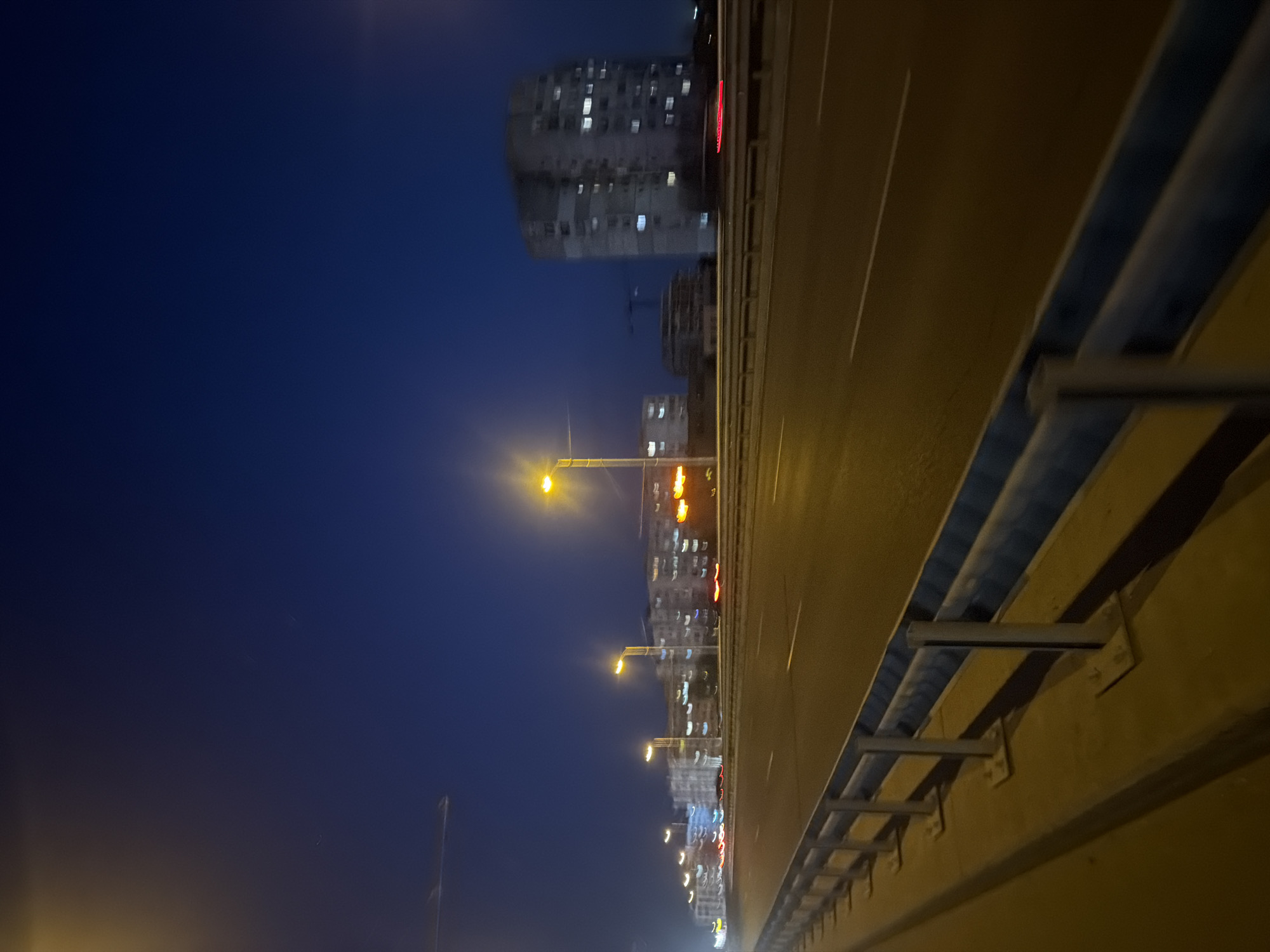 The image depicts a night scene in Sofia, Bulgaria, with a perspective from an elevated road or bridge. On the right side of the frame is the guardrail of the road. A dark blue night sky dominates the left side. Buildings are visible in the distance, illuminated with scattered lights. Streetlights are spaced along the road, casting a yellow glow. The asphalt appears dark and reflective.