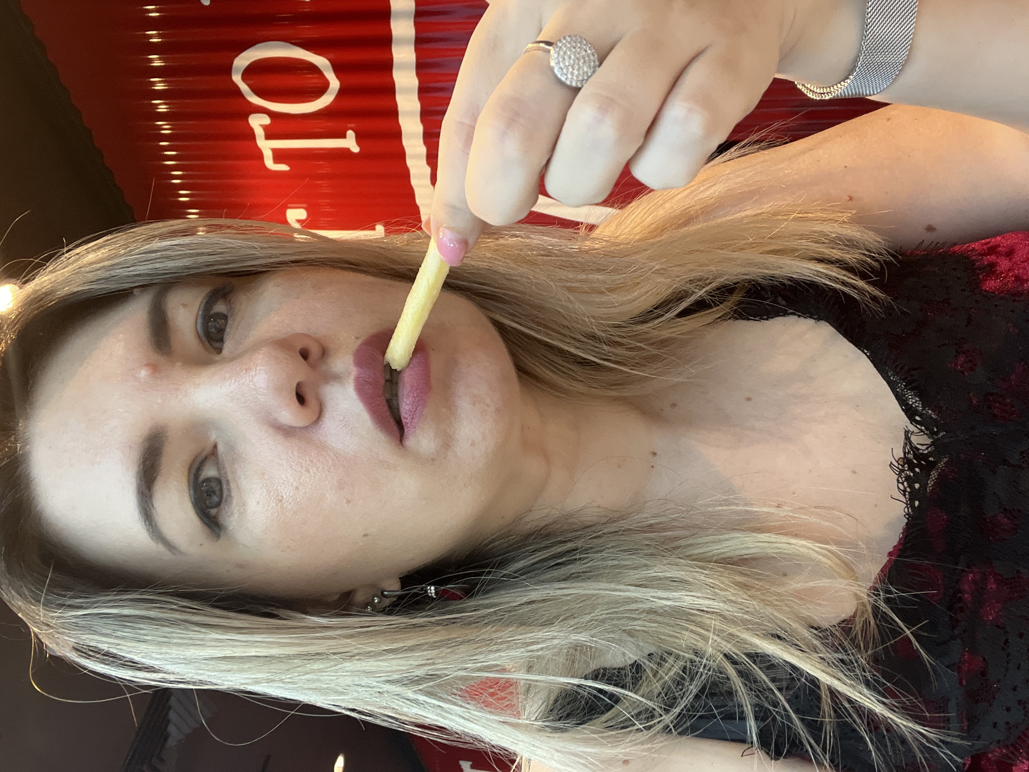 The image shows a close-up, rotated view of a woman eating a french fry. She has blonde hair and is wearing a black and red lace top. She is also wearing a sparkly ring on her right hand and a silver watch on her left wrist. The background is a red surface with white text that appears to be a logo or sign, with some of the letters visible. One of the visible words is "TO". The lighting suggests an indoor setting, possibly a restaurant, and the time of day is not discernible. The woman's expression is focused on the french fry she is eating.