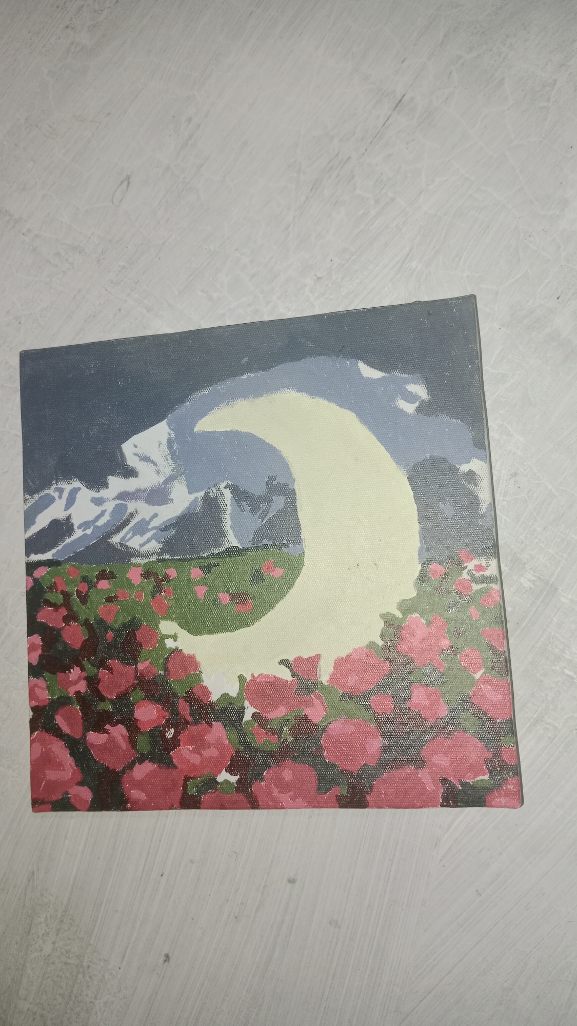 A painting on a canvas shows a crescent moon in a pale yellow hue, appearing in front of a backdrop of stylized mountains and a dark, cloudy sky. Below the moon, a field of pink roses with green foliage fills the lower portion of the image. The painting is placed on a light-colored, textured surface that appears to be a floor or table. There are no people or animals depicted in the painting. The painting does not have any identifiable landmarks or features that would indicate a specific location in Wono Sari, Indonesia.