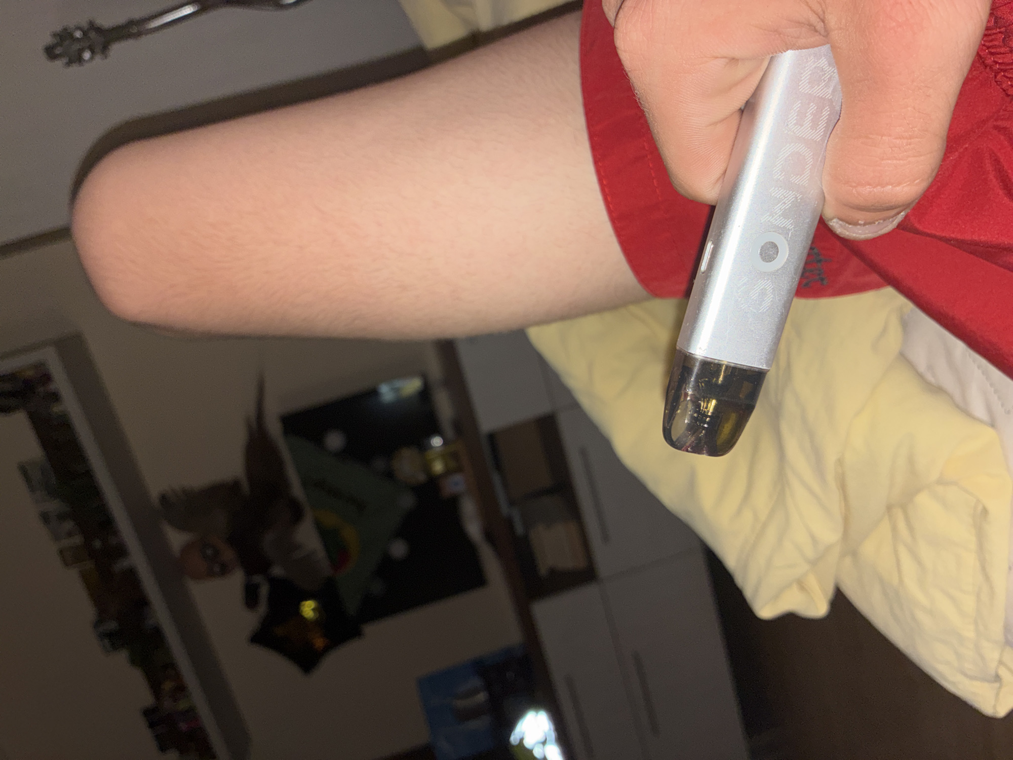 The image presents a close-up, angled view of a person's arm and hand holding a vaping device indoors. The arm, with fine hair visible, extends from the left side of the frame. The hand, with visible skin texture, firmly grasps a silver-grey cylindrical vaping device, possibly a pod system.
The device has a clear, dark-colored mouthpiece or pod at its lower end, through which some liquid is visible. The brand name "SONDER" is prominently displayed on the side of the device, along with a circular button or indicator. The person is wearing red fabric, likely shorts or pajamas, around their hand and arm, contrasting with a yellow duvet or blanket partially visible underneath.
The background is blurred, indicating an indoor setting, likely a bedroom or casual living space. Faintly visible on the left is a dark object, possibly a wall hanging or poster with some indistinct patterns. To the right, light-colored furniture, resembling cabinets or drawers, can be seen. A metallic, intricate object, possibly keys or a chain, is subtly present in the upper left corner.
The lighting appears subdued, suggesting an evening or night setting indoors. No specific activities are clearly depicted beyond holding the vaping device. No specific visual cues within the image indicate its location in Varna, Bulgaria.