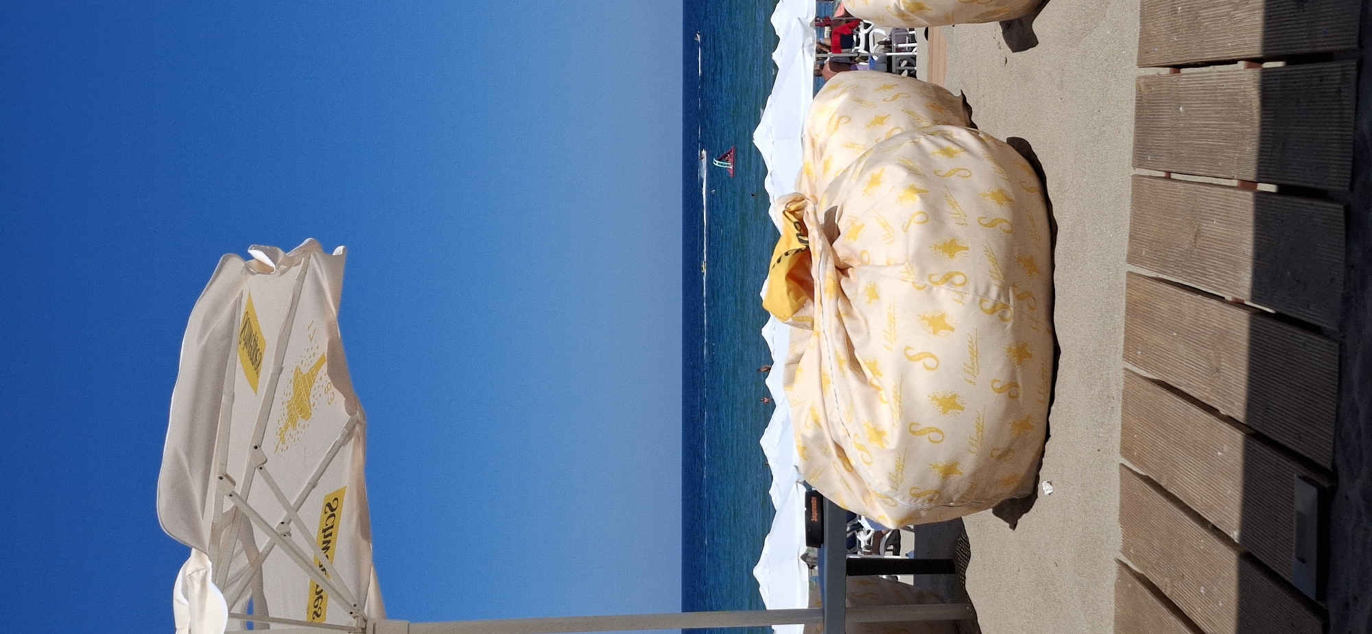 The image depicts a sunny day at a beach in Sozopol, Bulgaria. In the foreground, a wooden walkway is laid on the sand, leading towards the sea. Several large, round beanbag chairs with a light yellow fabric and a repeating pattern of yellow stars and the inscription "S. Hberger" are placed on the sand. One beanbag in the center is prominent, with a yellow top peeking out from its folds. Behind the beanbags, a row of white beach umbrellas extends towards the horizon. Further back, the blue sea stretches out, with a clear blue sky above. Several small boats and what appears to be a water toy are visible on the water. A few people can be seen in the distance, either on the beach or in the water. The overall atmosphere is one of a relaxed beach resort.