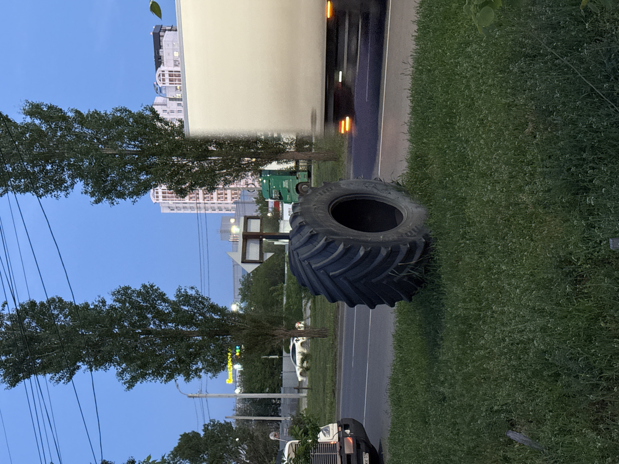The image shows a large tractor tire lying on a grassy shoulder adjacent to a paved road. A large white truck is partially visible, blurred due to movement, on the road next to the tire. In the background, several buildings are visible, including a high-rise apartment complex. A row of trees lines the roadside. A traffic light pole is also visible. Based on the available information, this photo was taken in the city of Ovoshchnoy, Russia.