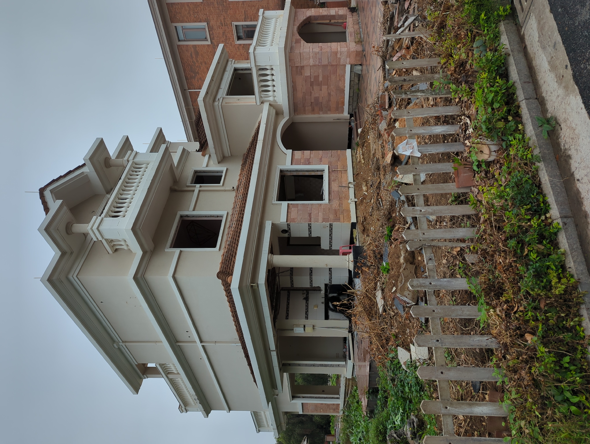 The image depicts an unfinished building in Shunde District, China. The building has a multi-story structure with exposed brick and concrete elements. The exterior lacks windows and doors in certain areas, revealing the interior. Debris and foliage surround the building, and a weathered wooden fence runs along the perimeter. The sky is overcast.