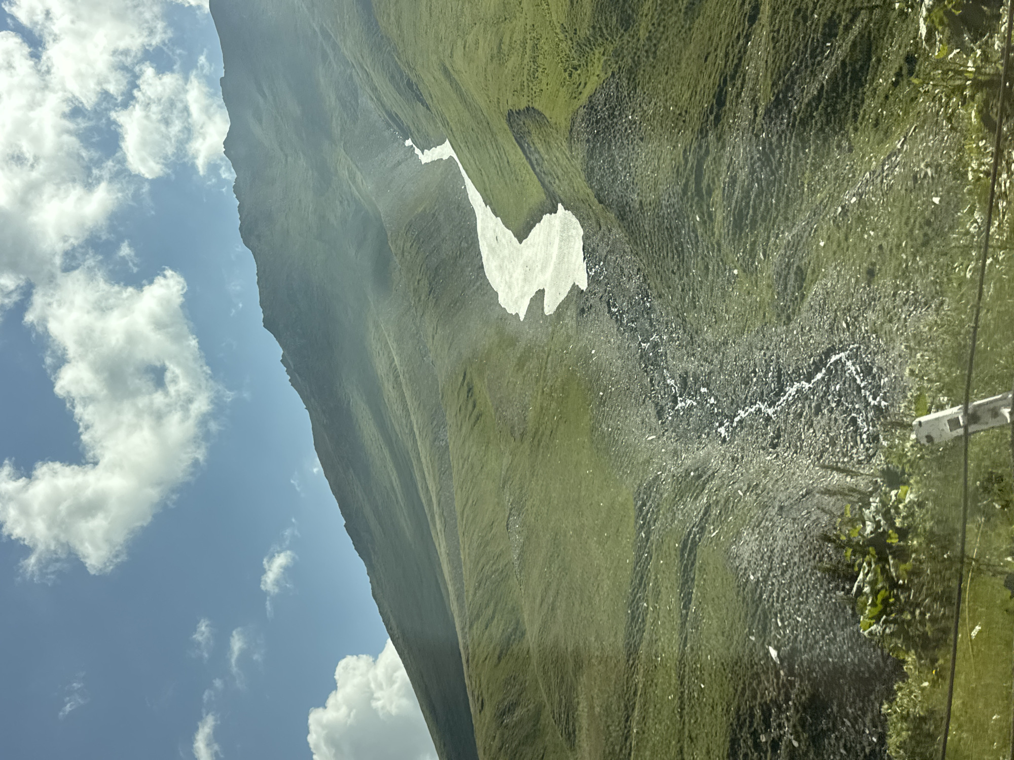 The image depicts a mountainous landscape with a clear blue sky dotted with white clouds. The slopes of the mountains are covered in lush green grass, with patches of rocky terrain. A winding path of snow or ice flows down the mountainside, forming a stream. The foreground shows some vegetation and a utility pole. The location is identified as Almasiani, Georgia.