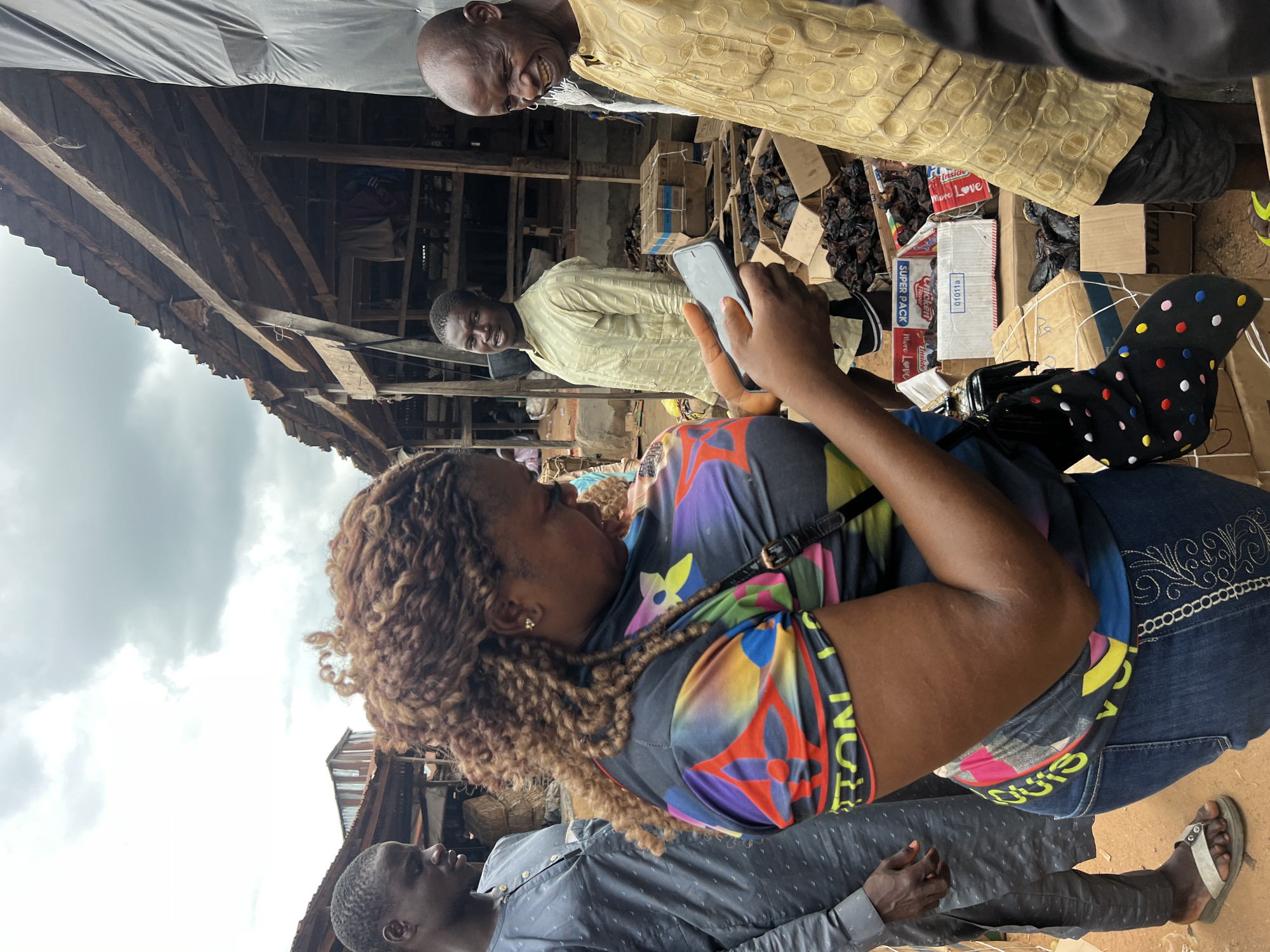 The image depicts an outdoor market scene in Asaba, Nigeria, under an overcast sky. In the foreground, a woman with braided brown hair is holding a smartphone and looking at it. She is wearing a colorful, patterned t-shirt and blue jeans. To her left, a man in a gray tunic stands looking towards the right. To her right, another man in a light yellow, patterned tunic is looking towards the camera. Further to the right, an older man with a bald head, wearing a yellow tunic, is looking towards the left and smiling broadly. The background reveals a bustling market with wooden stalls, some covered with dark tarpaulin. Several cardboard boxes are visible, some containing what appear to be dried goods. The ground is earthy. The atmosphere suggests a typical day at a Nigerian market, possibly a food market given the contents of the boxes.