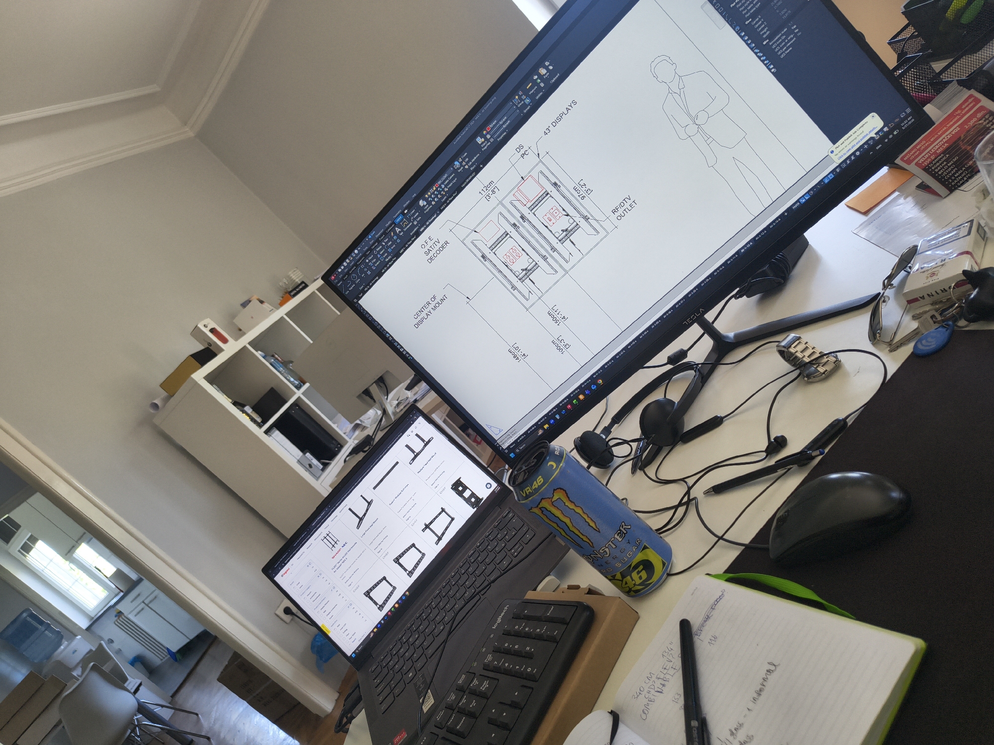 The image presents an indoor workspace. Two computer screens dominate the view. The larger screen displays technical drawings of displays and a man in a suit. The smaller laptop screen shows various schematic diagrams of brackets. In front of the laptops are a Logitech keyboard, a can of Monster Energy drink, a notebook, a pen, a mouse, and a pair of headphones. Other items on the desk include keys, sunglasses, and a pack of cigarettes. To the left is a white shelving unit with various objects. Through an open doorway, a view of another room with a window and additional furniture is visible. The OCR shows text in Serbian related to food items and prices, potentially indicating the user is in Belgrade, Serbia.