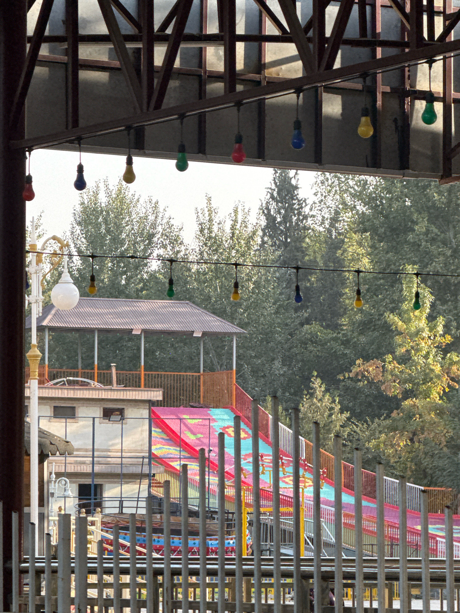 This is an outdoor daytime image captured in Dushanbe, Tajikistan, likely at an amusement park or recreational area. The foreground is dominated by a metal fence, beyond which is a colorful, multi-lane slide with red, pink, and turquoise sections, and yellow and orange railings. To the left of the slide is a multi-story building with a dark brown roof and a white facade. Above the slide and building, a canopy structure is visible, supported by dark brown beams. Several strings of colored light bulbs hang from the canopy, including red, blue, yellow, and green. A white lamp post with a round globe is also present to the left of the building. In the background, a dense forest of green trees fills the frame, with some autumn foliage visible. The sky is bright and overcast. No people are clearly visible.