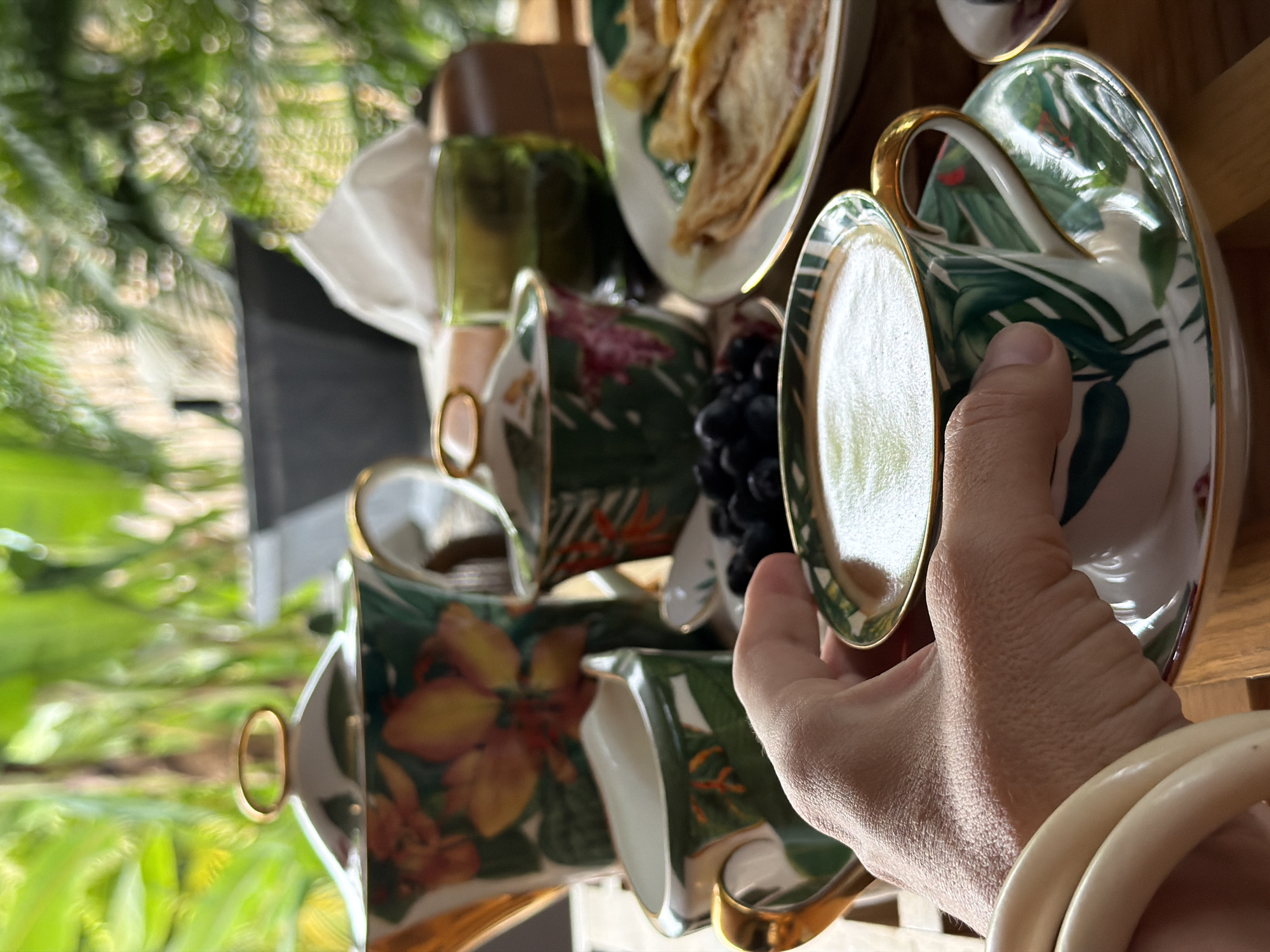 A close-up, eye-level shot shows a person's hand holding a coffee cup adorned with a tropical leaf pattern and gold trim. The cup rests on a saucer with a matching design. In the background, a breakfast spread includes a teapot, a sugar bowl, a creamer, blueberries, and pancakes. The scene is set outdoors, with lush green foliage visible, suggesting a tropical location. The overall impression is one of a tranquil breakfast enjoyed in a natural setting, possibly in Baan Bo Phut, Thailand.
