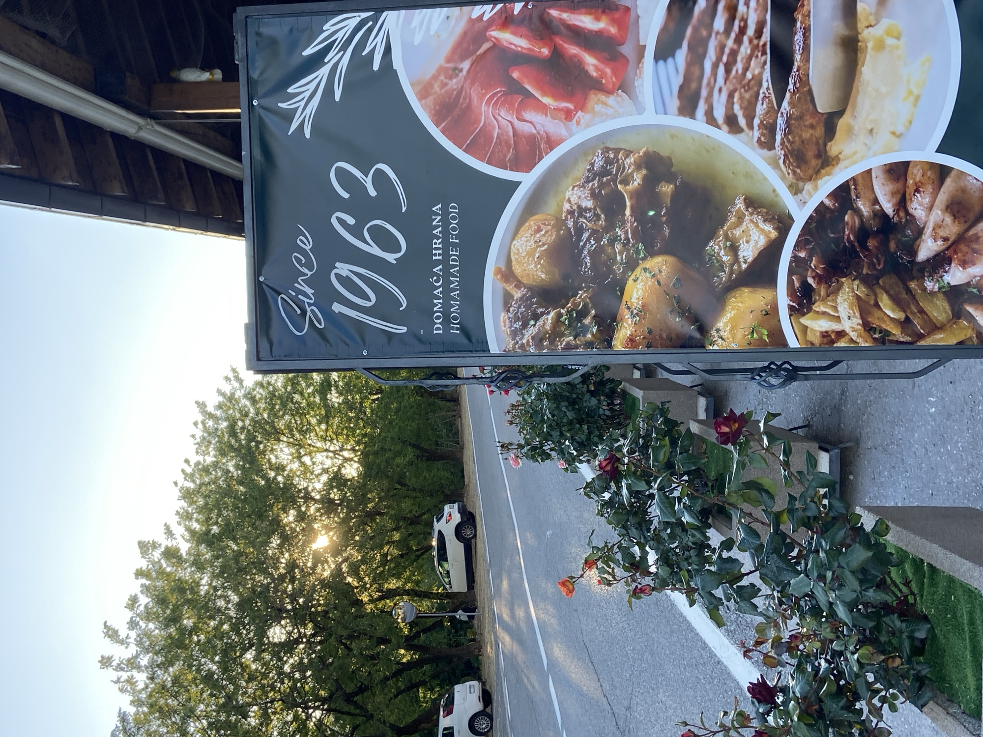 The image, taken in Sotonići, Montenegro, depicts an outdoor scene featuring a prominent banner for a food establishment. The banner, predominantly black with white text, displays "Since 1963" in a script font, followed by "DOMAĆA HRANA" and "HOMEMADE FOOD" in a sans-serif style. Circular insets on the banner showcase various dishes: slices of cured ham or prosciutto with tomatoes, grilled meats (possibly kebabs or ćevapi) served with mashed potatoes, a hearty stew with meat and potatoes, and another dish featuring sausages or grilled items with french fries.
Above the banner, on a wooden beam beneath a roof overhang, a white duck figurine is visible. In the foreground, a row of planters holds lush rose bushes with red and orange blossoms, bordering a paved road marked with white lines. Two white cars are parked alongside the road. The sun, low in the sky, shines brightly through the dense foliage of large green trees that line the left side of the scene, indicating late afternoon or early evening. The sky is clear, suggesting fair weather. No people are visible in the photograph.