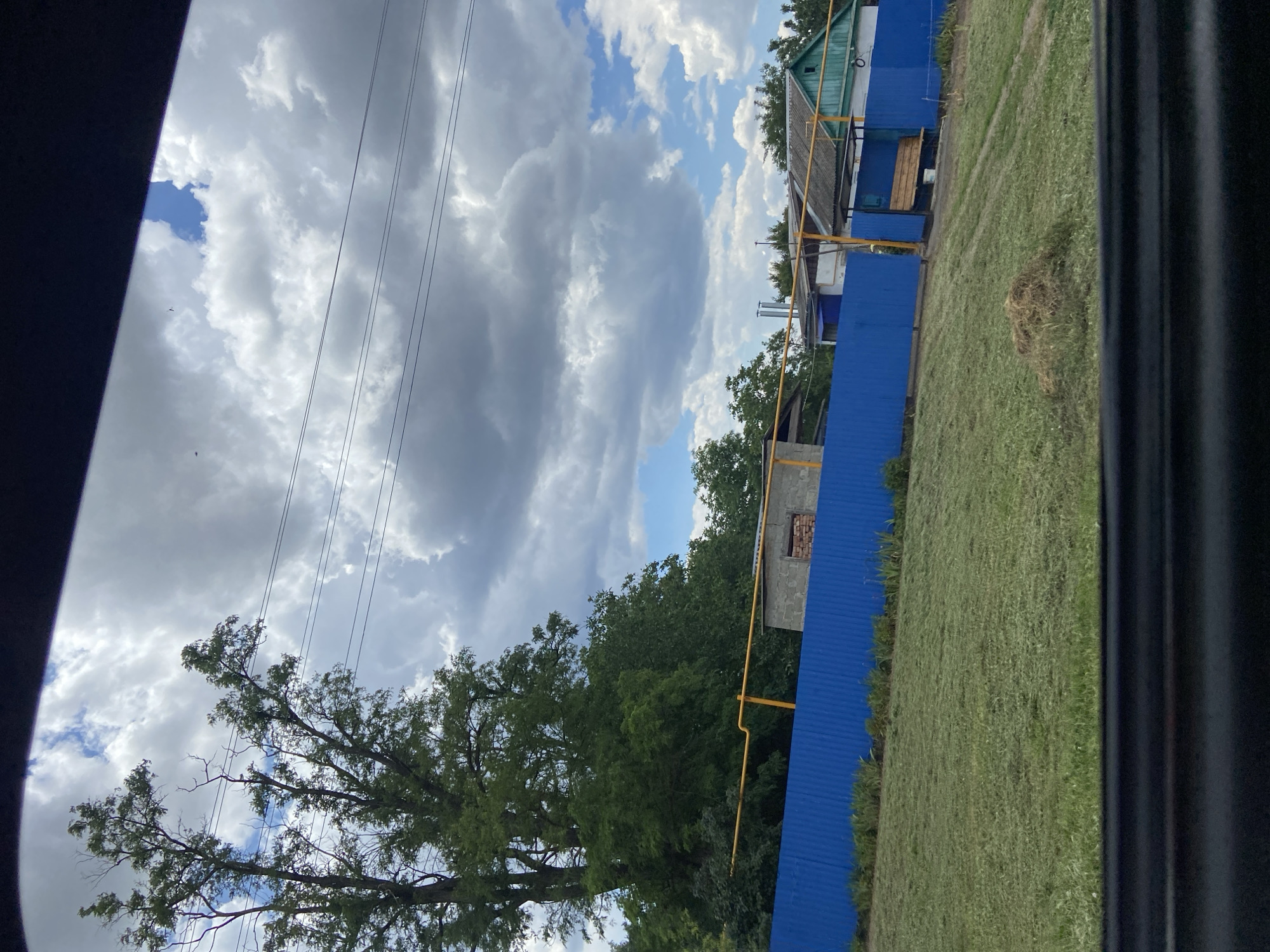 This is a vertical, wide shot of a rural outdoor scene in Taganrogsky, Russia. The sky is filled with a mix of fluffy white clouds and darker grey storm clouds, suggesting a dynamic weather system. Power lines stretch across the upper left of the frame.
Dominating the right side of the image is a bright blue corrugated metal fence. Beyond the fence, a neatly mown green lawn stretches out. In the distance, there are several structures, including a house with a teal roof and wooden porch, and other buildings partially obscured by trees. A yellow gas pipe runs horizontally along the fence line in several places. A pile of dried grass or straw sits on the lawn.
To the left of the fence, a large, leafy green tree with many branches extends upwards, its leaves rustling in the breeze. The overall impression is one of a slightly overcast day in a rural or suburban setting. There is no visible text in the image.