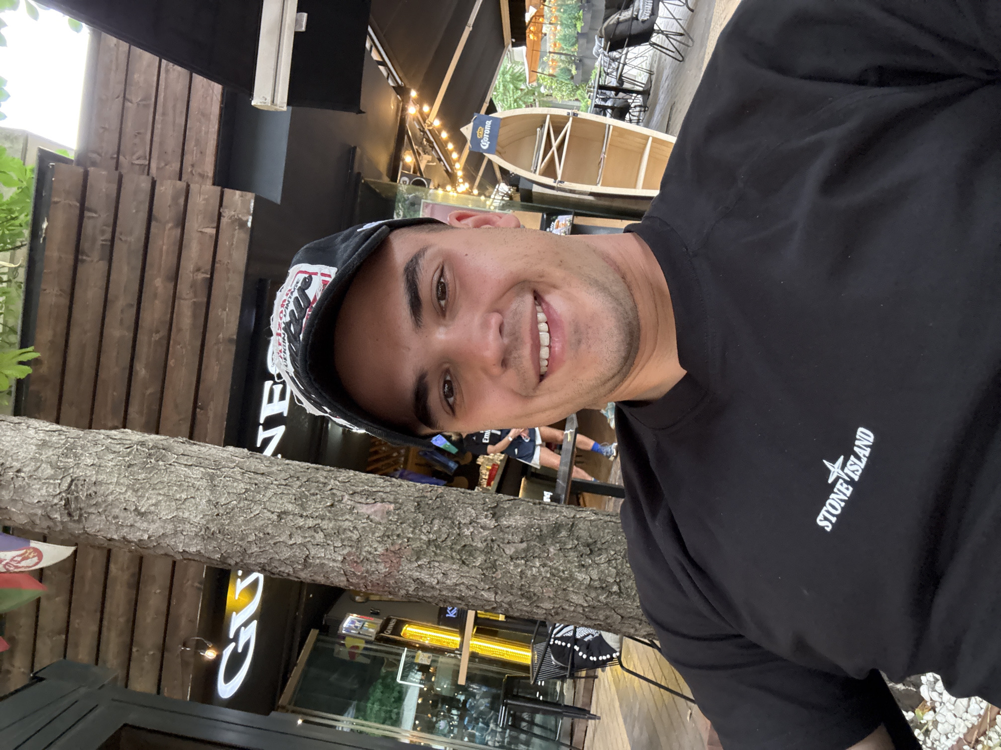A light-skinned male, estimated to be in his late 20s to early 30s, captures a direct selfie with a broad smile. He is wearing a black short-sleeved t-shirt featuring the white "STONE ISLAND" logo and compass rose emblem on the chest. A black baseball cap with a white and red graphic, possibly reading "ARIZONA VINTAGE," is worn backward on his head.
The setting is an outdoor area of a cafe or bar. A large, rough-barked tree trunk rises vertically through the mid-ground, partially dividing the scene. To the left of the tree, a glass-fronted building displays a white neon "GUINNESS" sign. A tall, yellow-orange radiant heater stands near black outdoor seating. In the upper-left corner, two small flags are visible, one clearly identifiable as the flag of Serbia.
To the right of the tree, the outdoor area features wooden decking, black outdoor tables and chairs, and a ceiling made of horizontal wooden slats. String lights are draped across the overhead structure. A distinctive wooden shelving unit, resembling a half-canoe or boat hull, stands upright, topped with a "Corona" branded sign. In the background, partially obscured individuals are visible, including one wearing a dark top with "Emirates" text and light-colored shorts. The environment suggests an urban cafe or restaurant setting in Serbia.