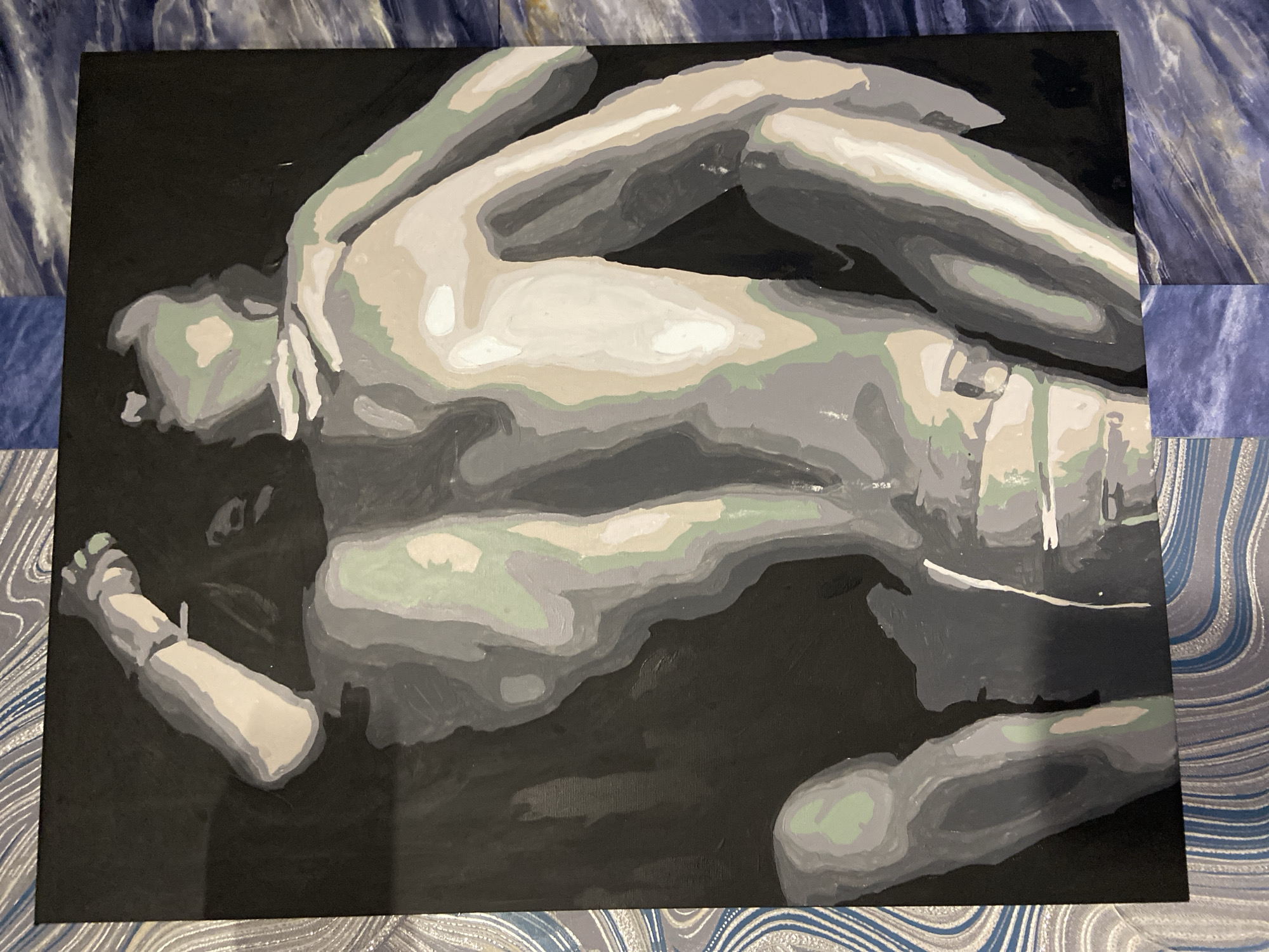The image depicts a painting of a nude figure rendered in a grayscale palette with hints of pale green and beige. The figure is shown from a high-angle perspective, lying on its back with limbs splayed. The painting style is abstract, with bold blocks of color defining the form and musculature. The background is a solid dark color. The painting is framed by a patterned material with blue and white swirls, which appears to be wallpaper or fabric. The overall composition is dark and dramatic, emphasizing the form of the body. There is no information present to determine the location of this painting within the city of Stavropol, Russia.
