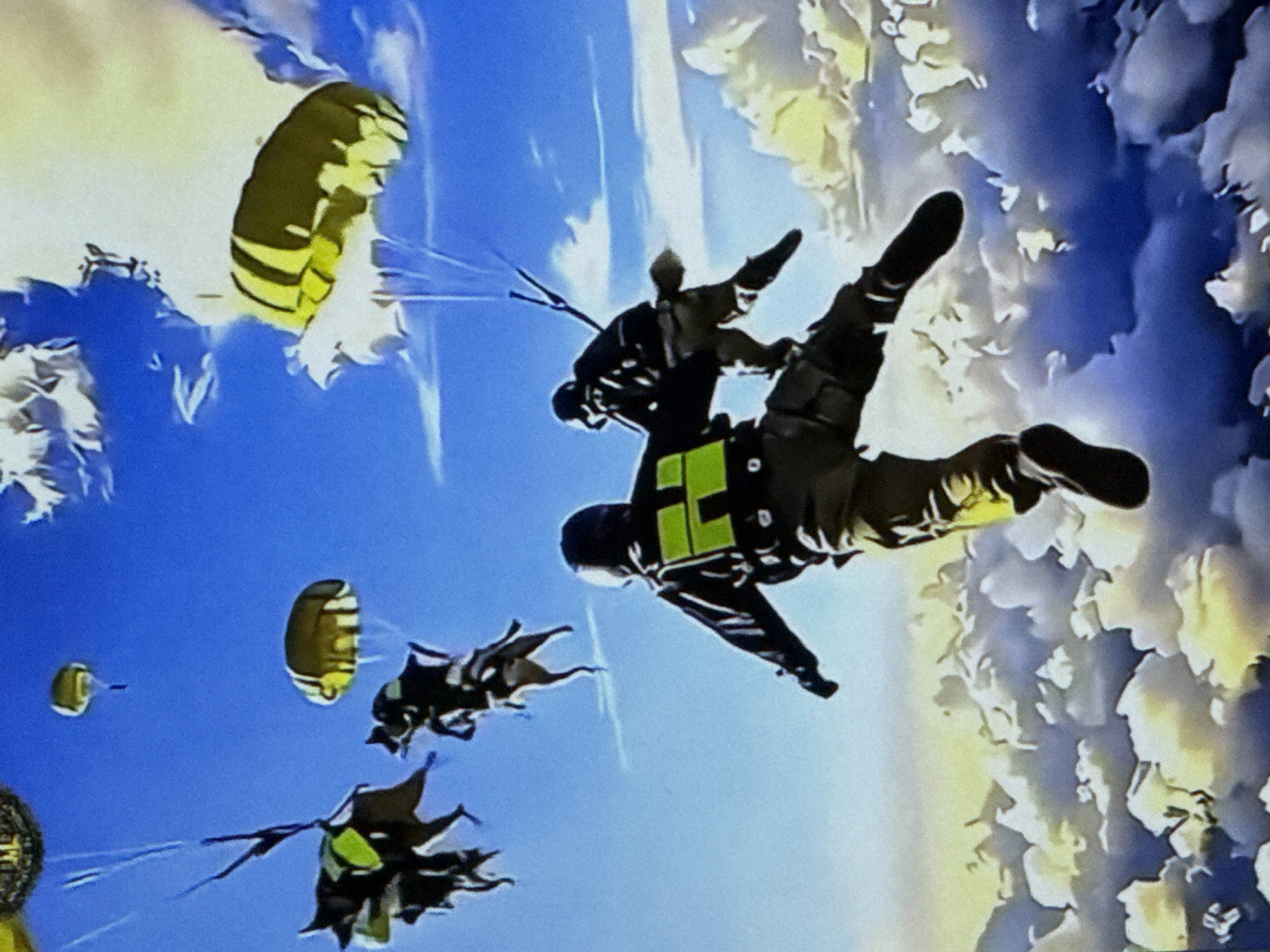 The media file shows a group of people skydiving against a backdrop of clouds and a bright blue sky, suggesting it is daytime. One prominent subject is a skydiver in the foreground, wearing a black suit with bright yellow-green accents and a helmet. They are in a freefall position, with their arms and legs extended. Another skydiver is visible further back, also in freefall. In the distance, several other figures and parachutes are present, some deployed and some still packed. The parachutes are brightly colored with yellow and black stripes. The scene is dynamic and exhilarating, capturing the thrill of skydiving. There is no visible text in the media file. The location is identified as Cahul, Moldova.