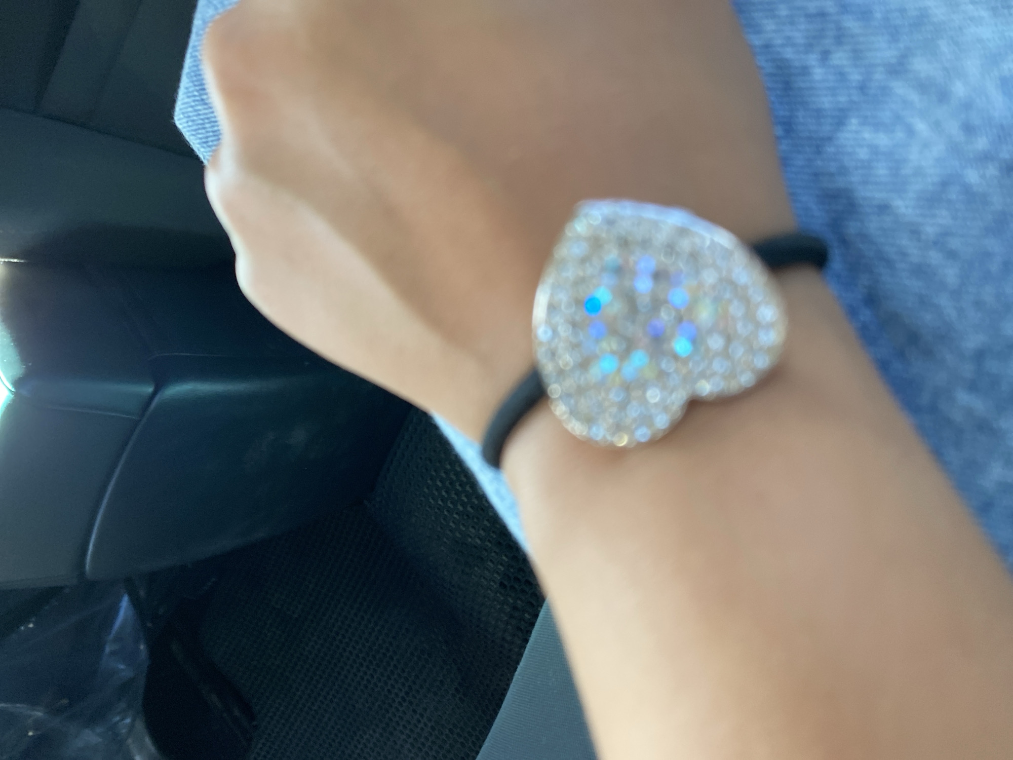 The image shows a close-up of a person's wrist wearing a black hair tie with a heart-shaped charm covered in rhinestones. The charm has iridescent blue and purple hues. The person is wearing a blue denim garment. The background is blurry but appears to be the interior of a vehicle, possibly a car, with dark seating and a mesh-like texture on what could be a floor mat. The specific location in Isyangulovo, Russia, cannot be determined from the visual information provided in the image.