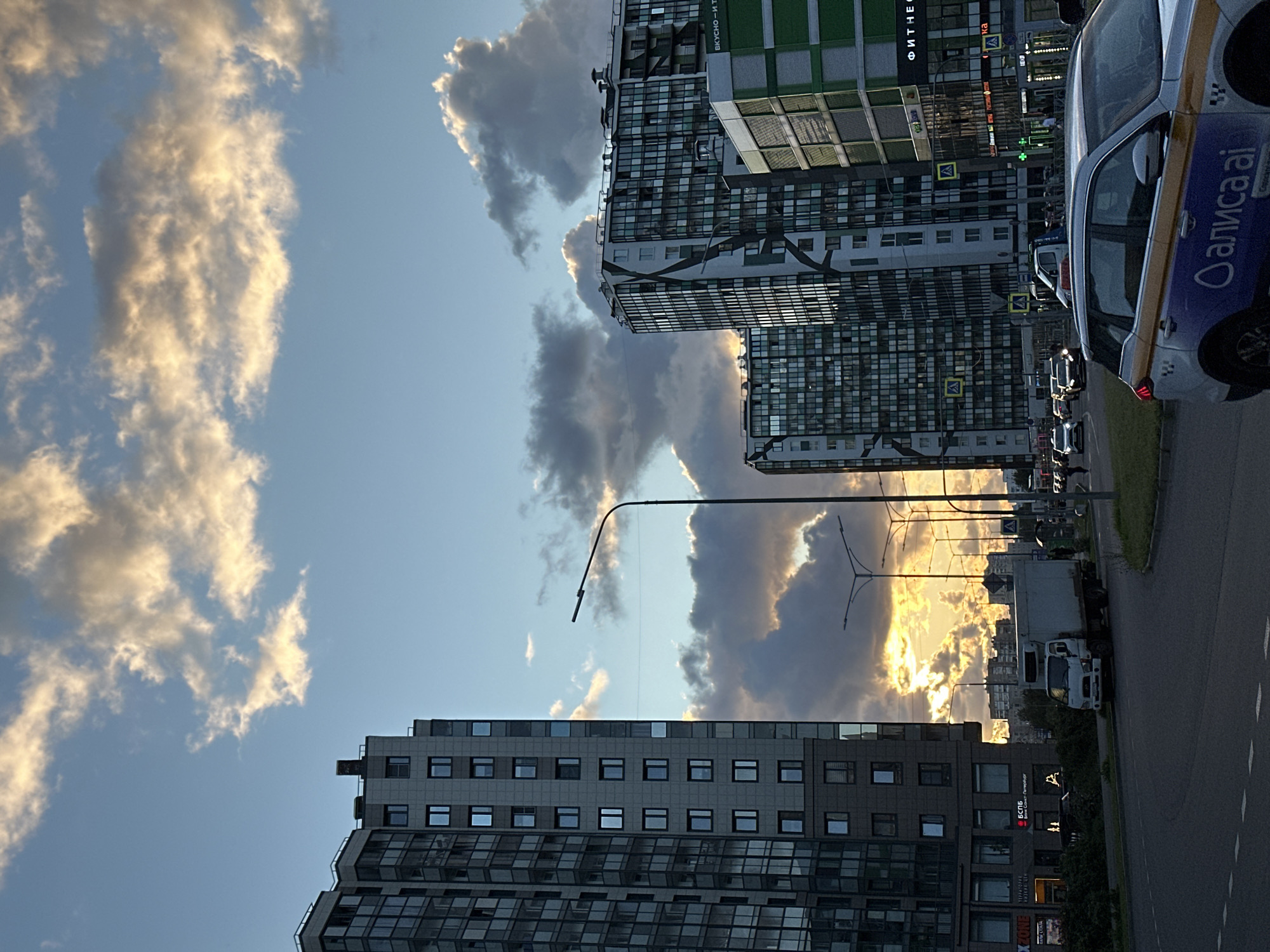 The image shows a street scene in Kudrovo, Russia, featuring modern apartment buildings, a cloudy sky illuminated by sunset, and traffic. In the foreground, a white car with blue and yellow taxi markings is parked. To the left, a tall residential building with many windows dominates the view. Across the street, several other apartment buildings are visible, some with a modern design featuring geometric patterns. A white truck is stopped on the road, and other cars are driving in the background. Streetlights and traffic signals are present. The sky is a mix of blue and dramatic clouds, with the setting sun casting a warm glow. The environment appears to be an urban setting with residential and commercial structures.
