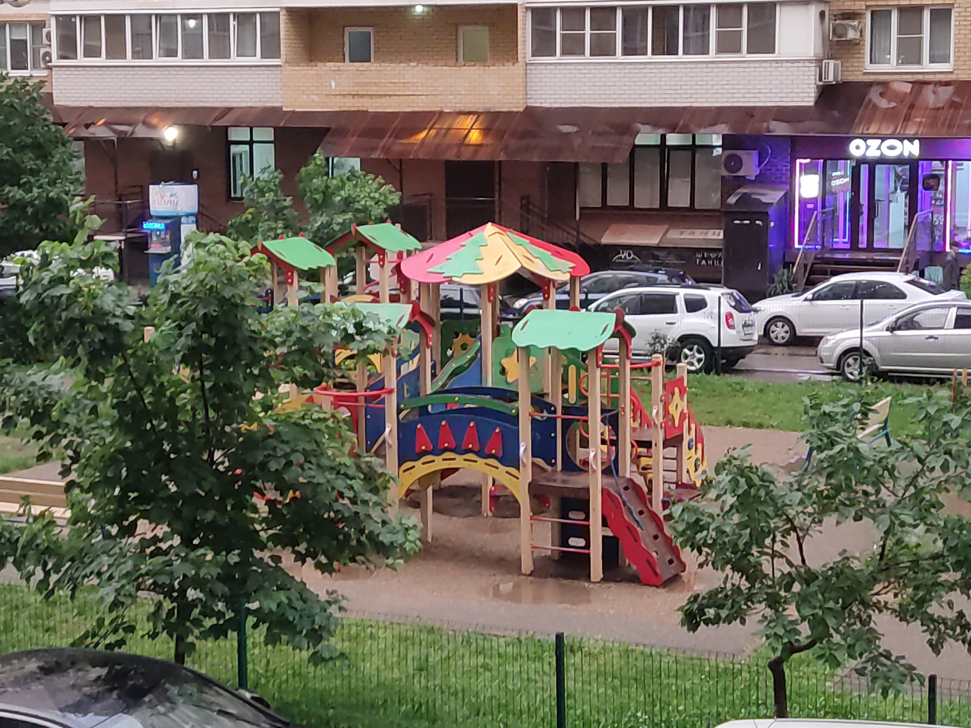 The image shows a playground in what appears to be a residential area. The playground features a multi-colored climbing structure with slides, bridges, and various play elements. Wet ground indicates it may have recently rained.
Behind the playground is a multi-story building with shops at ground level. One store has a sign that reads "OZON,". Cars are parked in front of the building. The location is Krasnodar, Russia based on the "OZON" store, which is a major Russian e-commerce company. The photograph seems to have been taken from an elevated position, possibly from a window in another building.