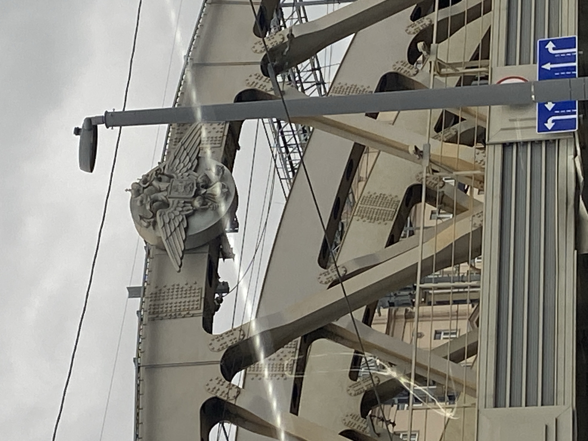 The image shows a close-up of the central span of the Bolshoy Moskvoretsky Bridge in Moscow, Russia, under an overcast sky. The bridge is constructed of beige-colored steel with numerous rivets visible on its structural elements. Prominently featured is a large, ornate coat of arms depicting two double-headed eagles, each crowned and with wings spread. The eagles are flanking a shield. To the right of the coat of arms, a grey lamppost with a modern, flat light fixture stands tall. The background reveals the skeletal structure of the bridge's supporting beams and a building with light-colored walls and windows. Below the bridge, a road surface with parallel grey and white stripes is visible, along with traffic signs indicating lane directions. The overall scene is static and industrial, with no people or vehicles clearly visible.