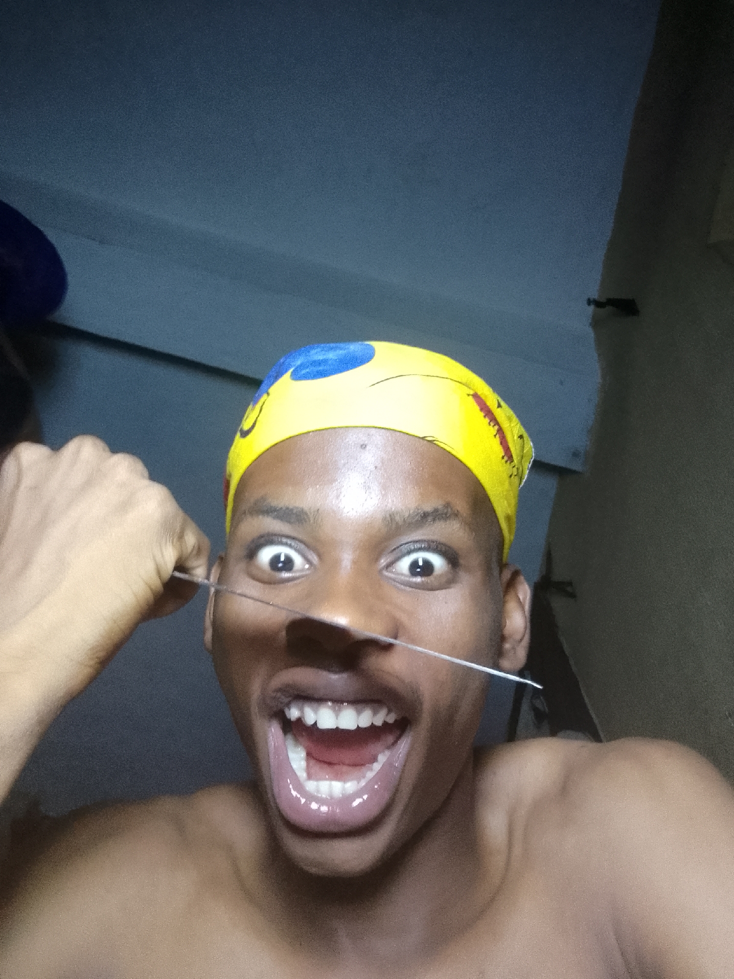 A dark-skinned person with a yellow and blue head covering is holding a thin metal object across their face, near the nose. The person's mouth is open wide with teeth showing. The location is inside a room in Nkpor, Nigeria, with gray walls.