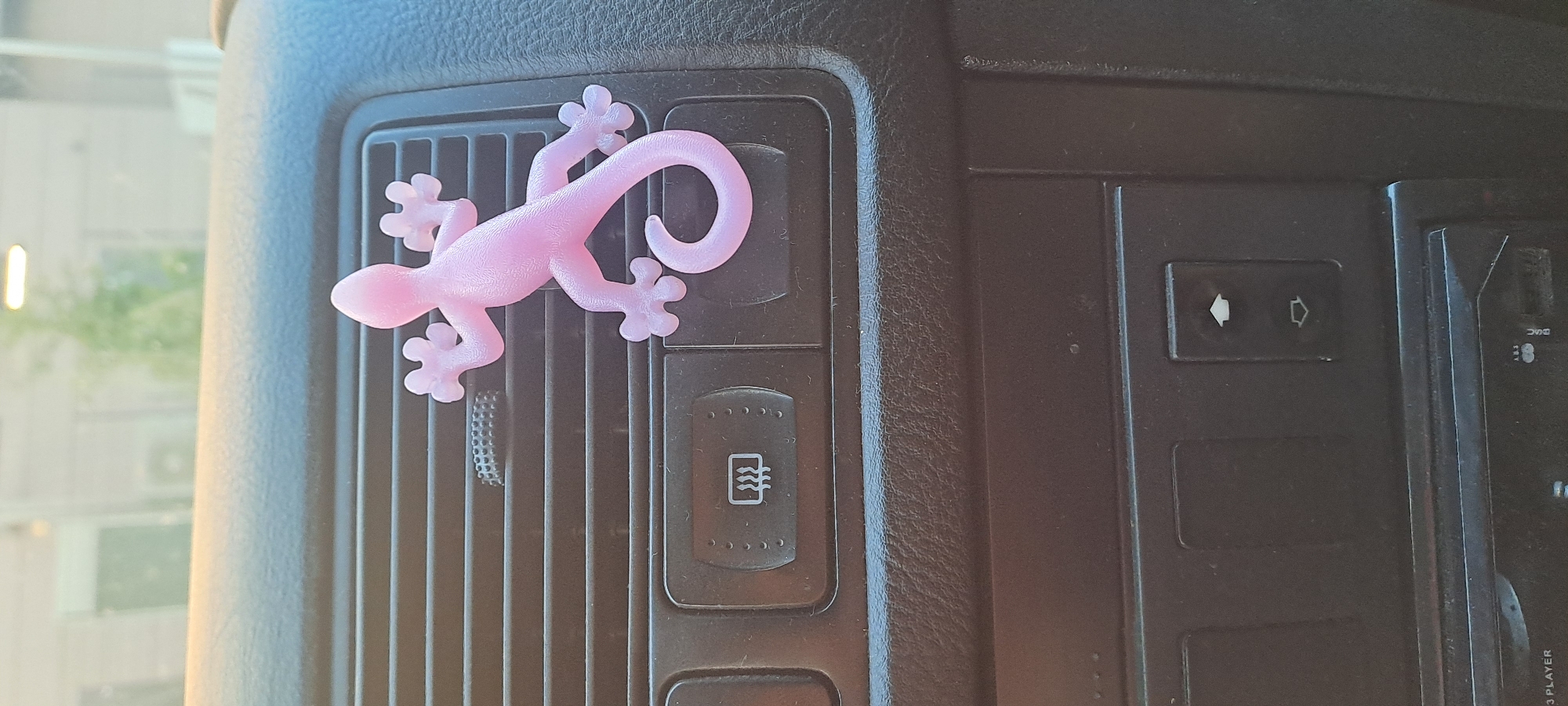 A bright pink, plastic gecko figurine is adhered to the horizontal slats of a car's air vent. The gecko is positioned on the right side of the air vent, with its body curved and its tail wrapped around. Below the air vent are two dark buttons. The left button has a white icon depicting wavy lines, possibly indicating a heated function. The right button has two white arrows, one pointing up and one pointing down. The dashboard surface is dark and textured. In the background, out of focus, is a blurred view of a building and greenery, suggesting an outdoor environment. The lighting suggests daylight.