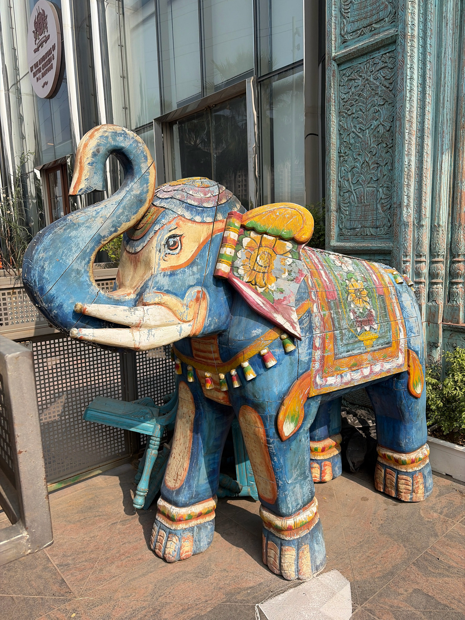 This image captures a vibrant, weathered, wooden elephant statue situated outdoors in Gurgaon, India. The statue, predominantly painted in hues of blue, yellow, and orange, is adorned with intricate, colorful patterns on its back, trunk, and legs, suggesting a decorative Indian motif. The elephant's tusks are a chipped, off-white color, and its ears are a faded blue. A small, turquoise-colored stool with decorative carvings sits behind the elephant's front left leg.
The background features a modern building with a glass facade, reflecting sunlight and the surrounding environment. A circular sign with text and a logo is visible on the building's exterior. To the right of the elephant, an elaborately carved, distressed turquoise wooden pillar adds to the richly decorative, possibly South Asian-inspired, aesthetic of the scene. Green foliage is visible in the lower right corner, indicating a planted area. The ground is paved with textured, reddish-brown tiles. The overall atmosphere is bright and sunny, with clear shadows indicating daytime. The scene appears to be a public or commercial space, likely an outdoor seating area or decorative entrance.