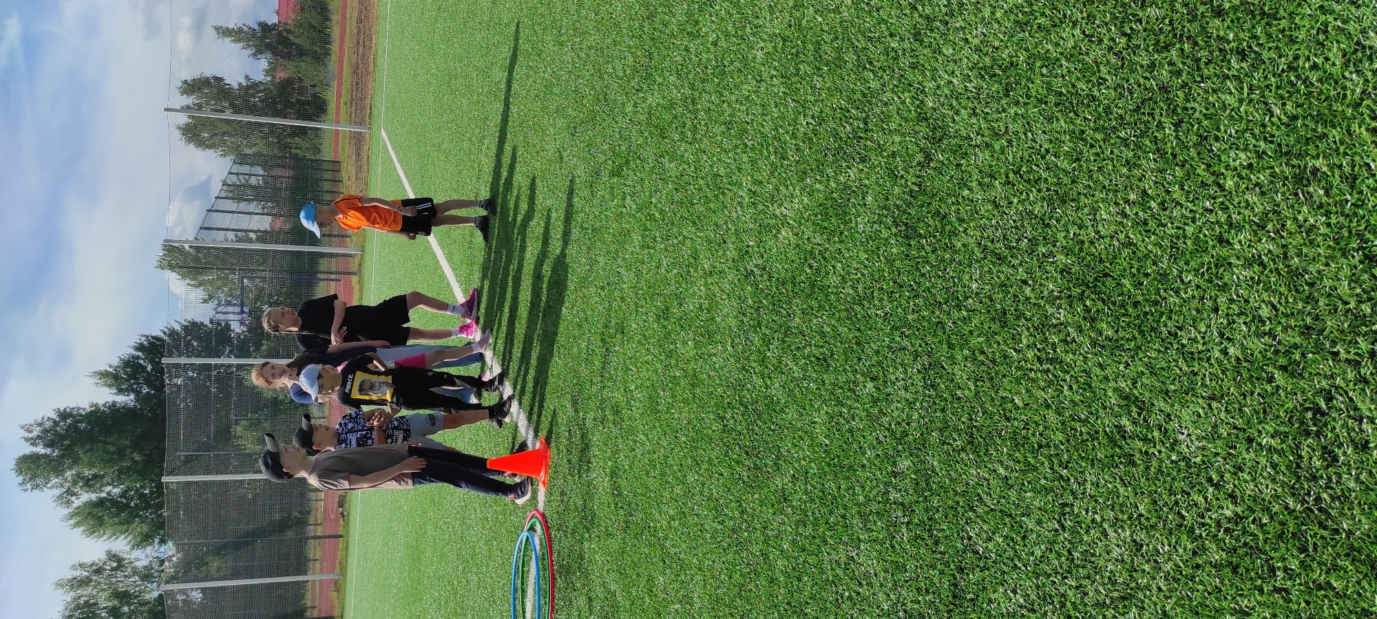 A group of five children, appearing to be between 8 and 12 years old, are standing on a bright green artificial turf field, likely a sports field, in Noviy, Russia. The field is marked with white lines. The children are dressed in casual summer clothing. One boy in an orange t-shirt and black shorts stands slightly apart from the others, facing them. To the left of the group, several colorful hula hoops are lying on the ground, next to an orange traffic cone. In the background, a chain-link fence encloses the field, and beyond that, trees and some buildings are visible under a cloudy sky. The sun casts distinct shadows on the grass, suggesting it is daytime. The overall scene appears to be an outdoor recreational activity or sports practice.