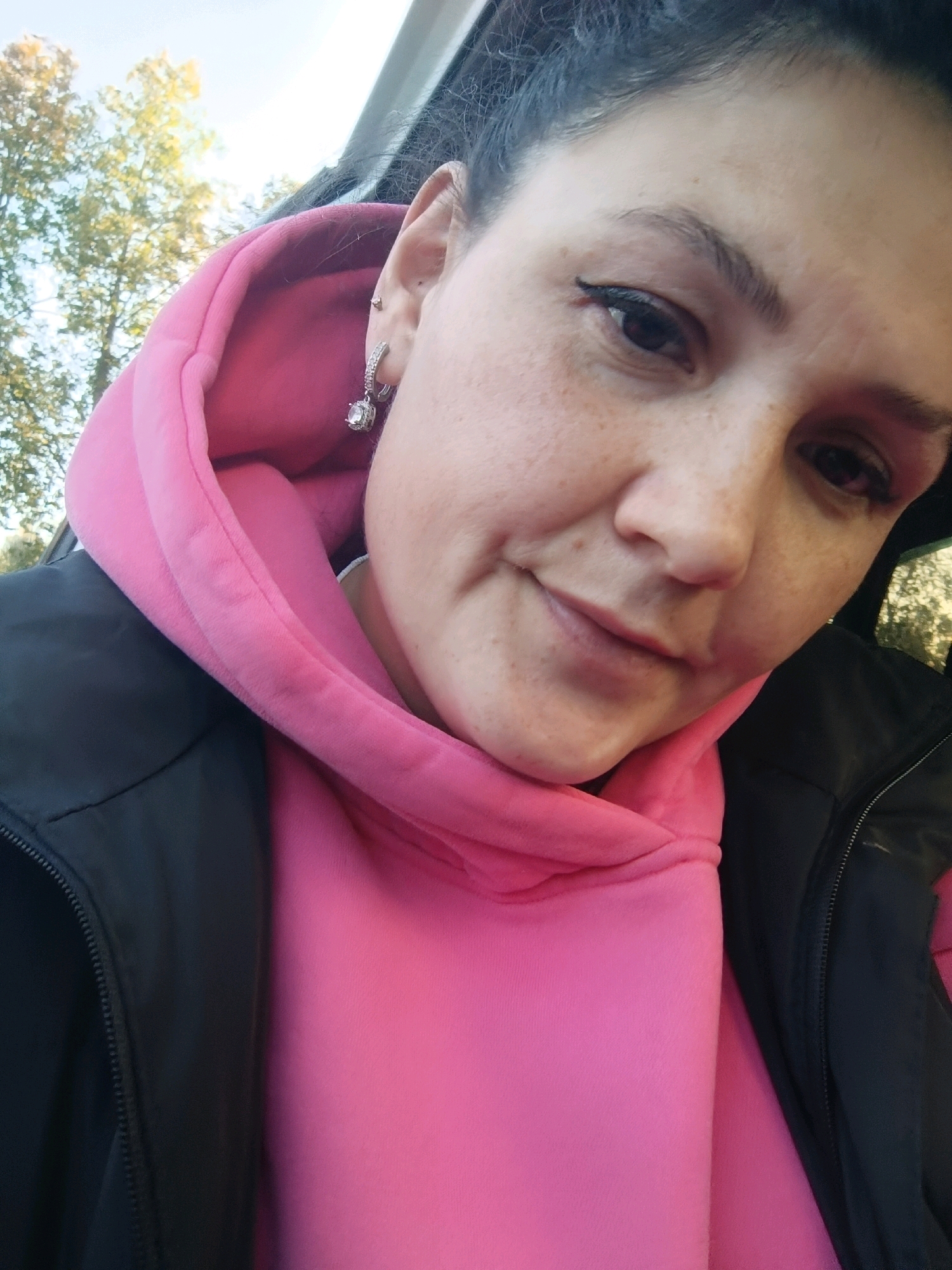 A woman is taking a selfie in a car, likely parked in the city of Chaikovsky, Russia, as suggested by the background foliage. The woman has dark hair pulled back, a pink hoodie, and a black jacket. She is wearing a silver hoop earring with a clear stone and has eyeliner and freckles visible. The lighting suggests it is daytime.