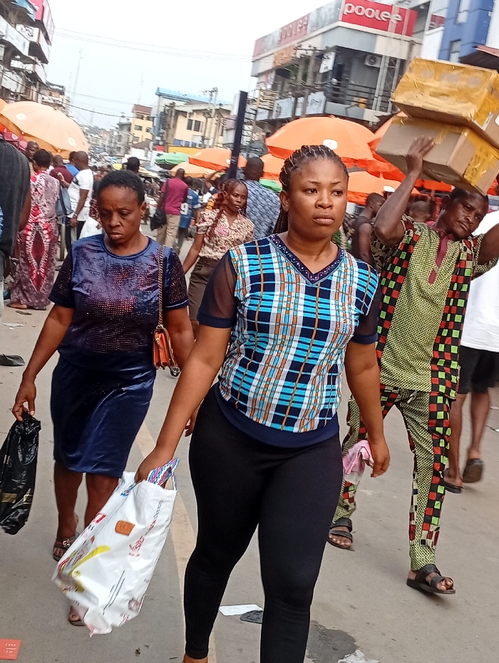 A bustling street scene shows two women walking in the foreground. The woman on the right wears a blue and white checkered shirt with black leggings, carrying a white shopping bag. The woman to her left wears a blue dress and carries a black plastic bag. A man carrying boxes on his head in a patterned shirt walks to their right. The background is filled with people, umbrellas, and buildings, creating a vibrant and crowded market atmosphere.