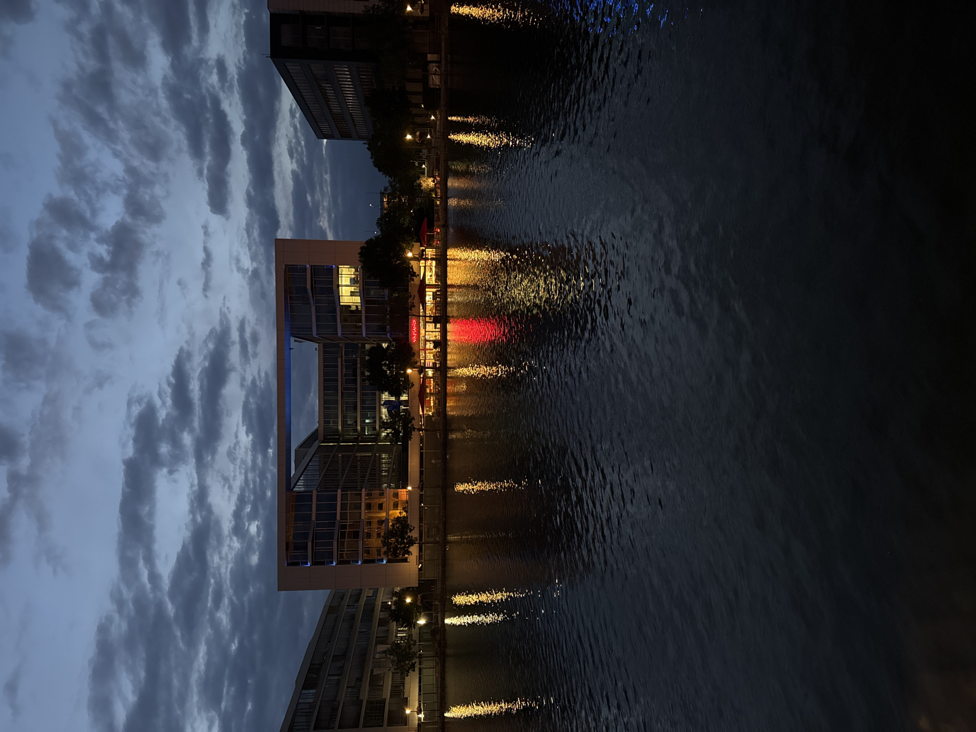 This image shows a modern office building with a large rectangular opening in the center, reflected in the dark water of a canal or river. The building is illuminated from within, with some lights on, and a restaurant with red awnings is visible at the base of the building. The sky is dark blue with scattered clouds, suggesting it is dusk or early evening. The reflection of the building and lights creates streaks of color on the water's surface. This scene is located at the Portsmouth-Platz in Duisburg, Germany.