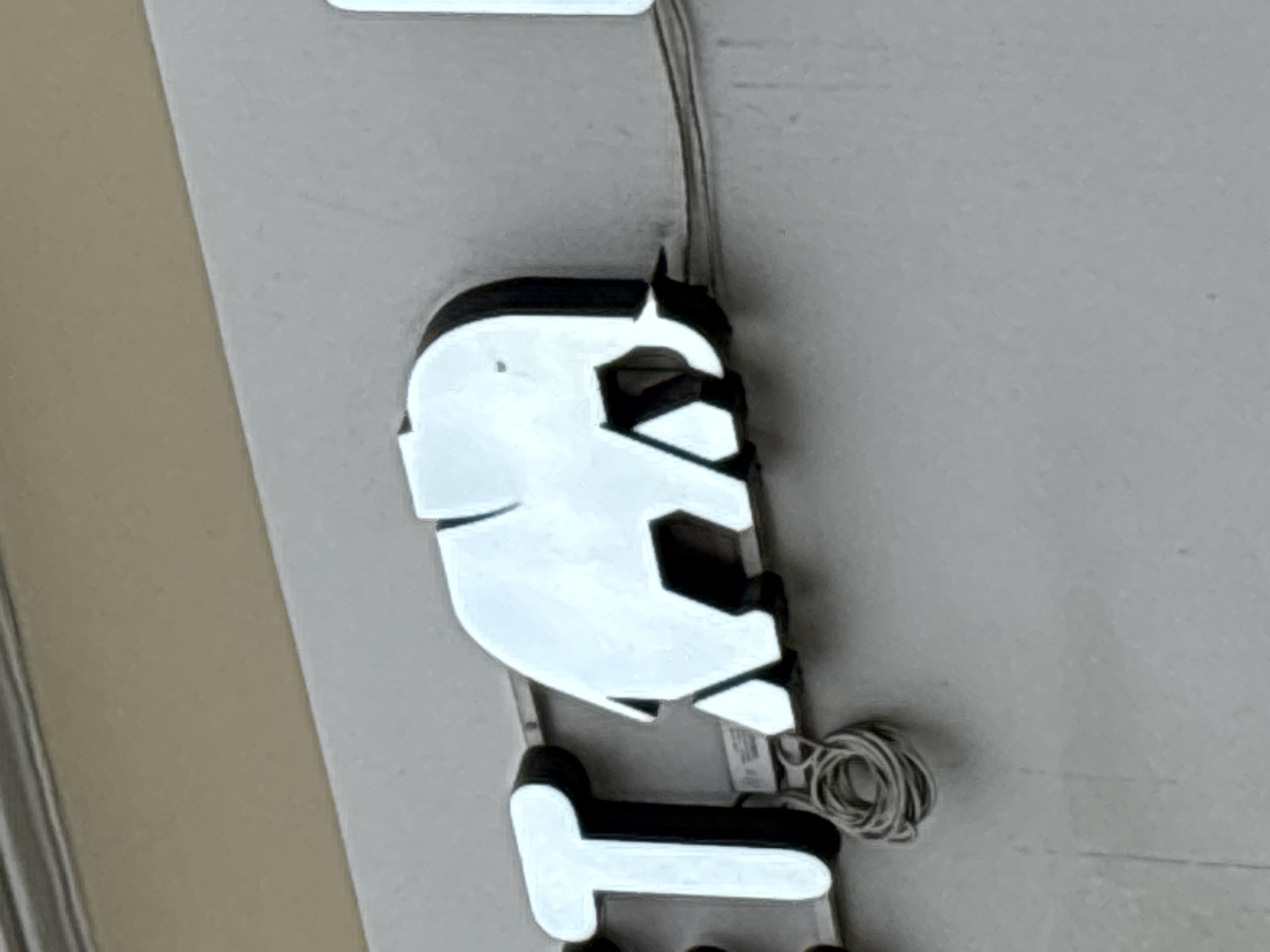 A white elephant silhouette logo is mounted on a grey wall. The logo is illuminated from behind and appears to be made of metal. The elephant is facing to the right and is in a walking pose. A coiled wire is visible below the elephant. The logo is part of a sign for the "Elephant" art space located at 18 Nevsky Prospekt, Saint Petersburg, Russia.