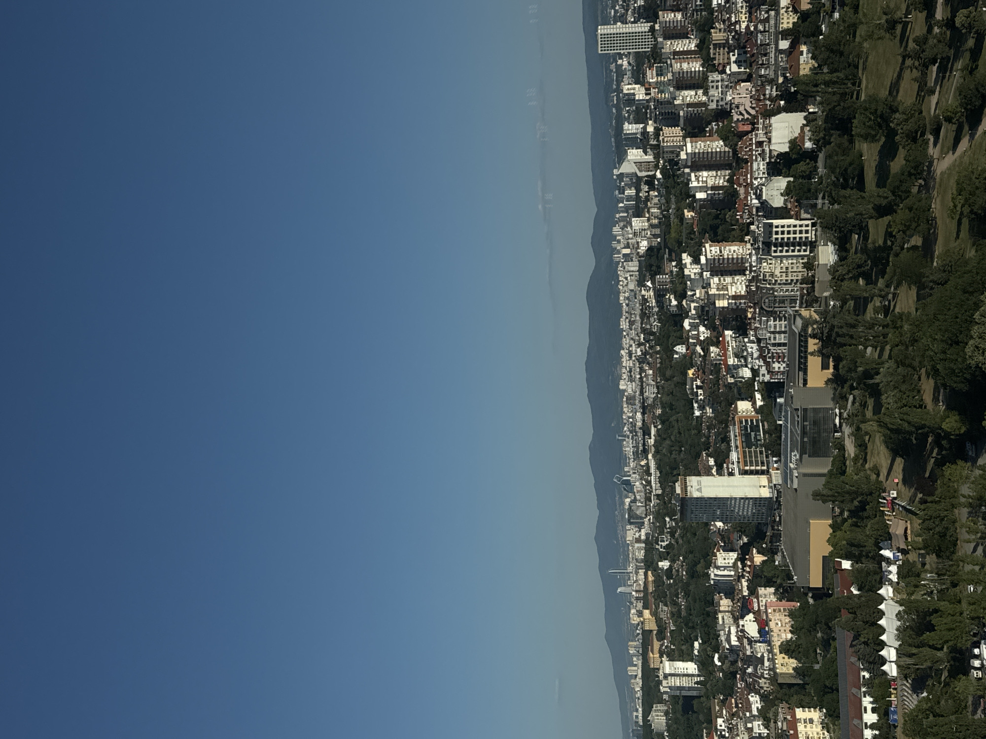 This image is an aerial view of Sofia, Bulgaria, looking east. In the foreground, a park area features a stage with white tents and a few scattered people. Beyond the park, a dense urban landscape unfolds, characterized by a mix of residential and commercial buildings, including a large yellow building complex that appears to be an exhibition center. The midground is dominated by numerous apartment buildings and office structures of varying heights and architectural styles. In the distance, a mountain range forms the horizon under a clear blue sky. Several prominent modern skyscrapers are visible within the city, suggesting a mix of older and newer developments. The overall impression is of a sprawling city nestled against a natural backdrop.