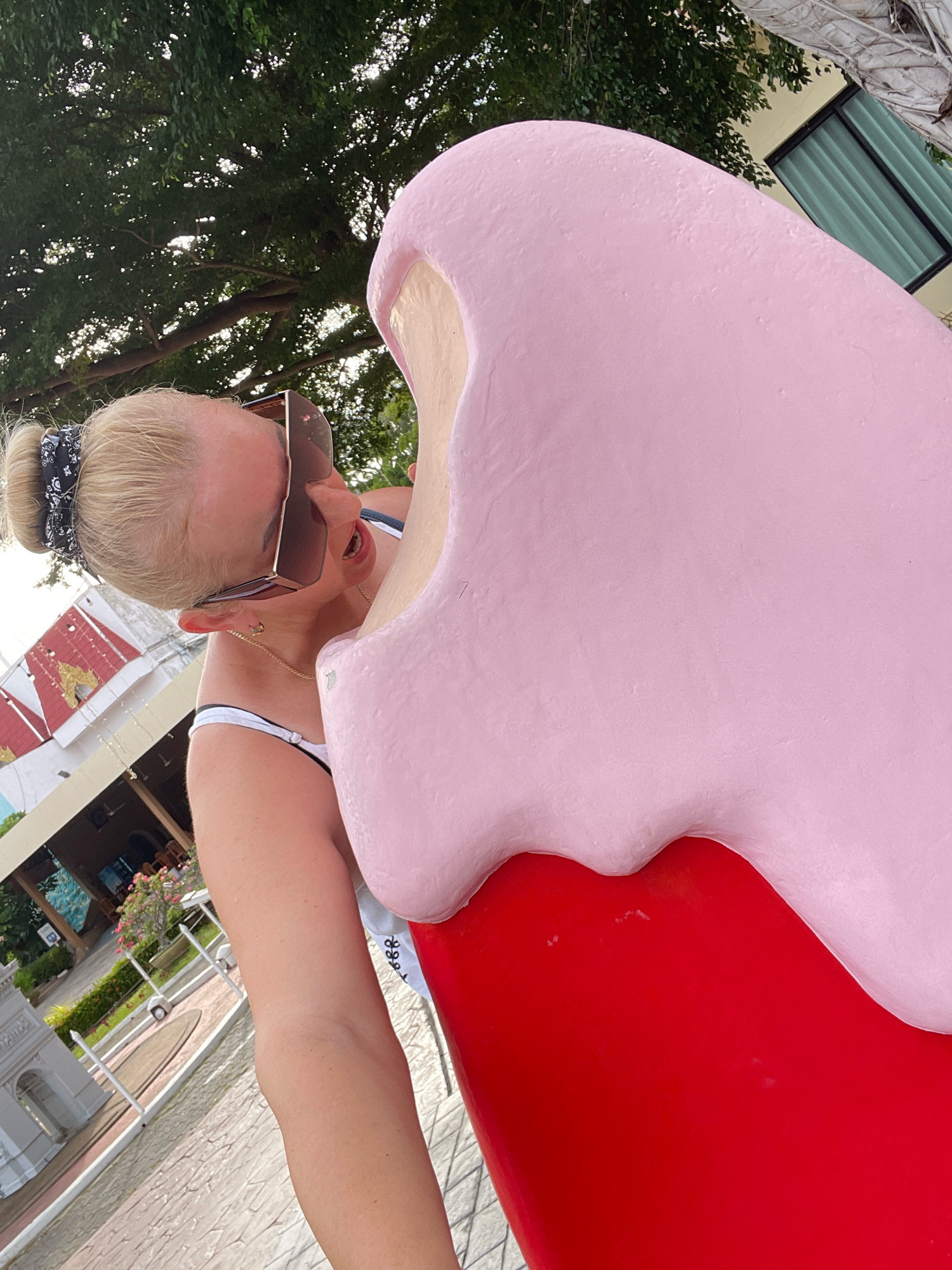 A blonde woman with a bandana in her hair and large sunglasses is posing for a selfie next to a giant pink ice cream cone sculpture. The sculpture is red at the base and pink on top. She is wearing a bikini and is leaning towards the sculpture. In the background, there are trees, a building with a red roof, and a window with green curtains. The scene appears to be outdoors, possibly in a park or a tourist attraction. The location is Pattaya City, Thailand.