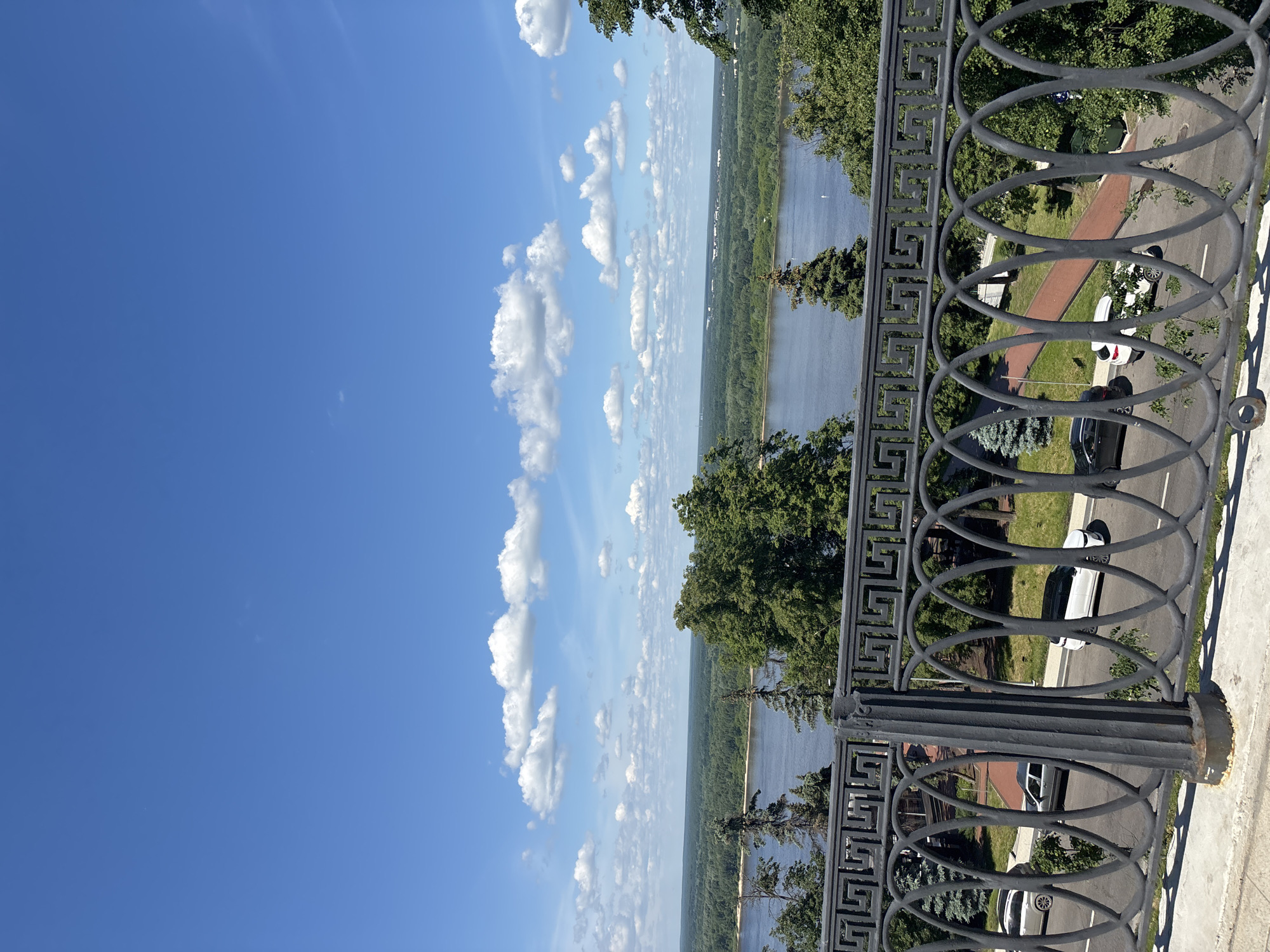 The image captures a panoramic landscape view from an elevated vantage point overlooking a wide river, likely the Volga, in Nizhny Novgorod, Russia. The scene is depicted on a clear, bright day under a vivid blue sky dotted with scattered white cumulus clouds.
In the foreground, a dark metal decorative railing runs horizontally across the bottom of the frame, suggesting a balcony or observation deck. The railing features intricate patterns, including intertwined circles and a geometric Greek key-like motif. Peeking through and below the railing, one can see a paved road or drive with several parked cars, including a white SUV and a dark sedan. Beyond the road, there's a grassy area with some trees and a reddish-brown path, indicating a park or landscaped area descending towards the river.
The midground is dominated by the expansive river, its surface reflecting the bright sky. The opposite bank is covered by a dense, verdant forest stretching towards the horizon, with a narrow strip of sand or beach visible in parts along the water's edge. Some trees are also visible closer to the railing, framing the view.
The time of day appears to be midday or early afternoon, characterized by the strong, natural light and the clear sky. The weather is sunny and pleasant. There is no visible text in the image. The overall impression is one of serene natural beauty combined with an urban riverside vista.