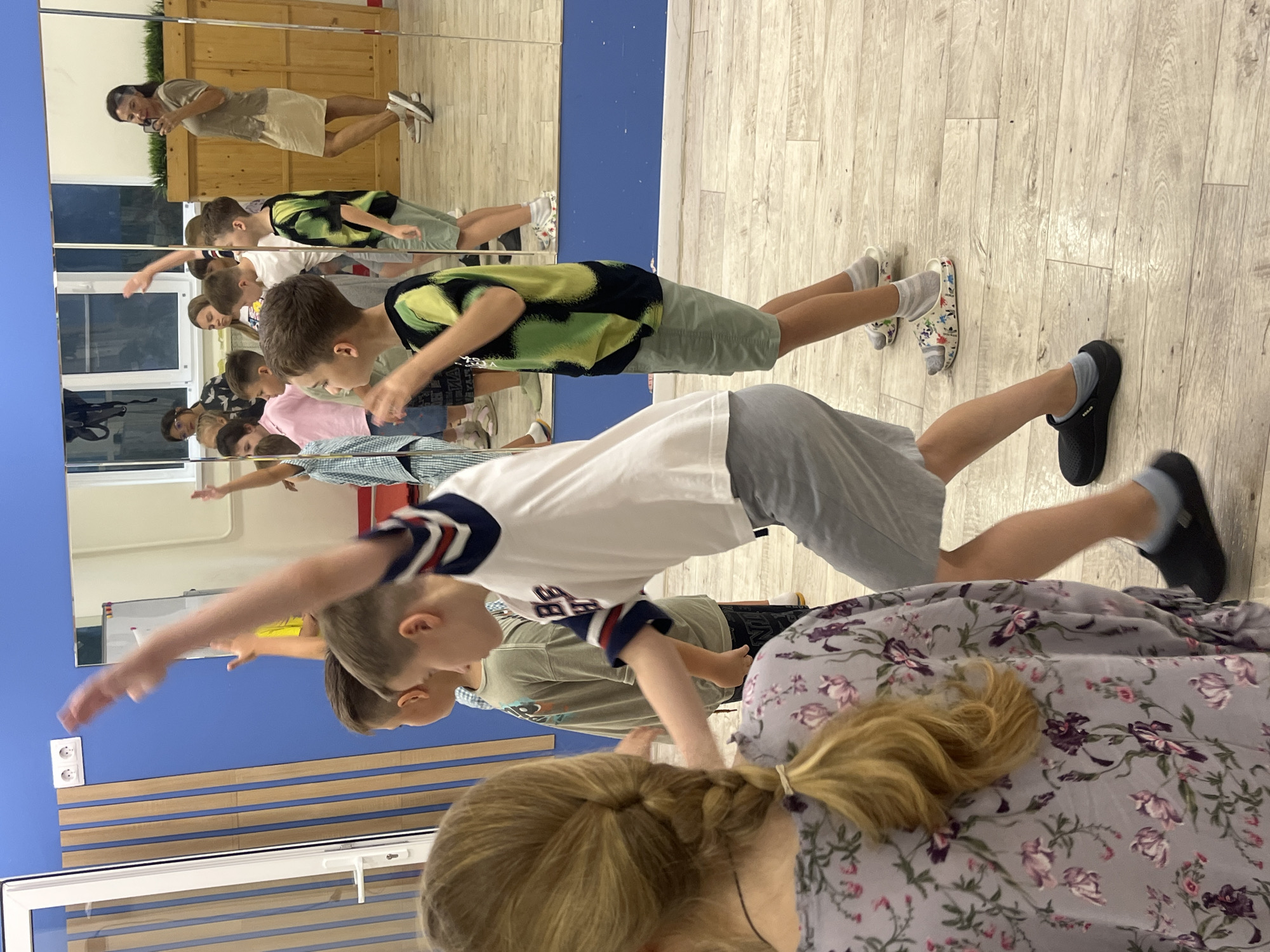 The image depicts an indoor dance class or activity session for children, with a woman, likely an instructor, visible in the background through a mirror. The room appears to be a studio or gym, featuring a mirrored wall, a blue painted wall, and wooden flooring. Several children are actively engaged in what seems to be a dance or exercise routine, with their arms raised and bodies in motion.
The children are dressed casually in t-shirts and shorts. One boy in the foreground is wearing a white t-shirt with navy and red stripes on the sleeves and grey shorts. Another boy to his right is in a black and green tie-dye t-shirt and light green shorts. Behind them, other children are partially visible, some in patterned shirts and shorts.
In the reflection of the mirror, a woman with dark hair, wearing a light brown top and a beige skirt, is holding a phone, possibly recording the class. She is standing in front of a wooden panelled wall with some greenery. The overall impression is one of an active and engaging session for the children. The specific location in Sochi, Russia, cannot be determined from the visual information provided in the image.