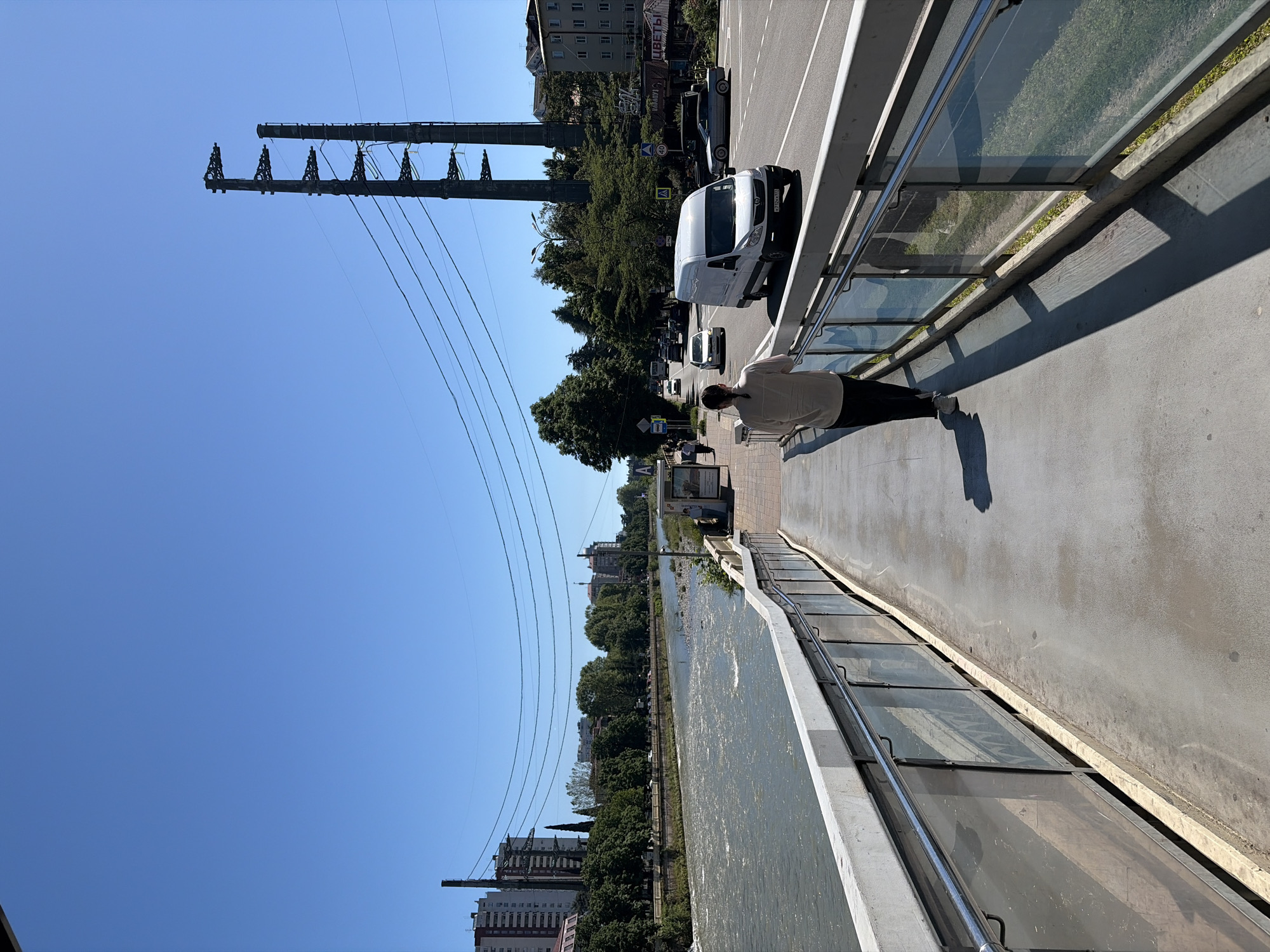 The image depicts a bright, sunny day in Sochi, Russia, captured from an elevated pedestrian walkway or bridge. The scene overlooks a river, a busy road, and urban infrastructure.
In the foreground, a person wearing a light-colored shirt and dark pants stands on the concrete walkway, leaning against a low wall that features a glass barrier, looking towards the river. The walkway has reflections on its surface from the bright sunlight. Shadows are cast strongly, indicating midday.
To the right of the walkway, a road runs parallel, with several vehicles, including a prominent white van (license plate "E072XX 93") and other cars, suggesting active traffic. Along the road and riverbank, dense green trees line the landscape, providing a lush contrast to the concrete and asphalt. Behind the trees, various buildings, including multi-story apartment blocks, are visible.
A significant feature in the midground and background are large, robust power line structures or towers with multiple parallel lines stretching across the sky. These industrial elements stand out against the clear, vibrant blue sky that dominates the upper portion of the image. The river, with slightly rippled water, flows alongside the road and beneath the power lines. On the far bank of the river, more buildings and trees extend into the distance.
Visible text includes a sign or banner above a shop on the roadside displaying "ЦВЕТЫ" (TSVETY), which is Russian for "FLOWERS." Another partial sign with "А" followed by unreadable text is also present. The clear sky and bright conditions suggest pleasant weather.