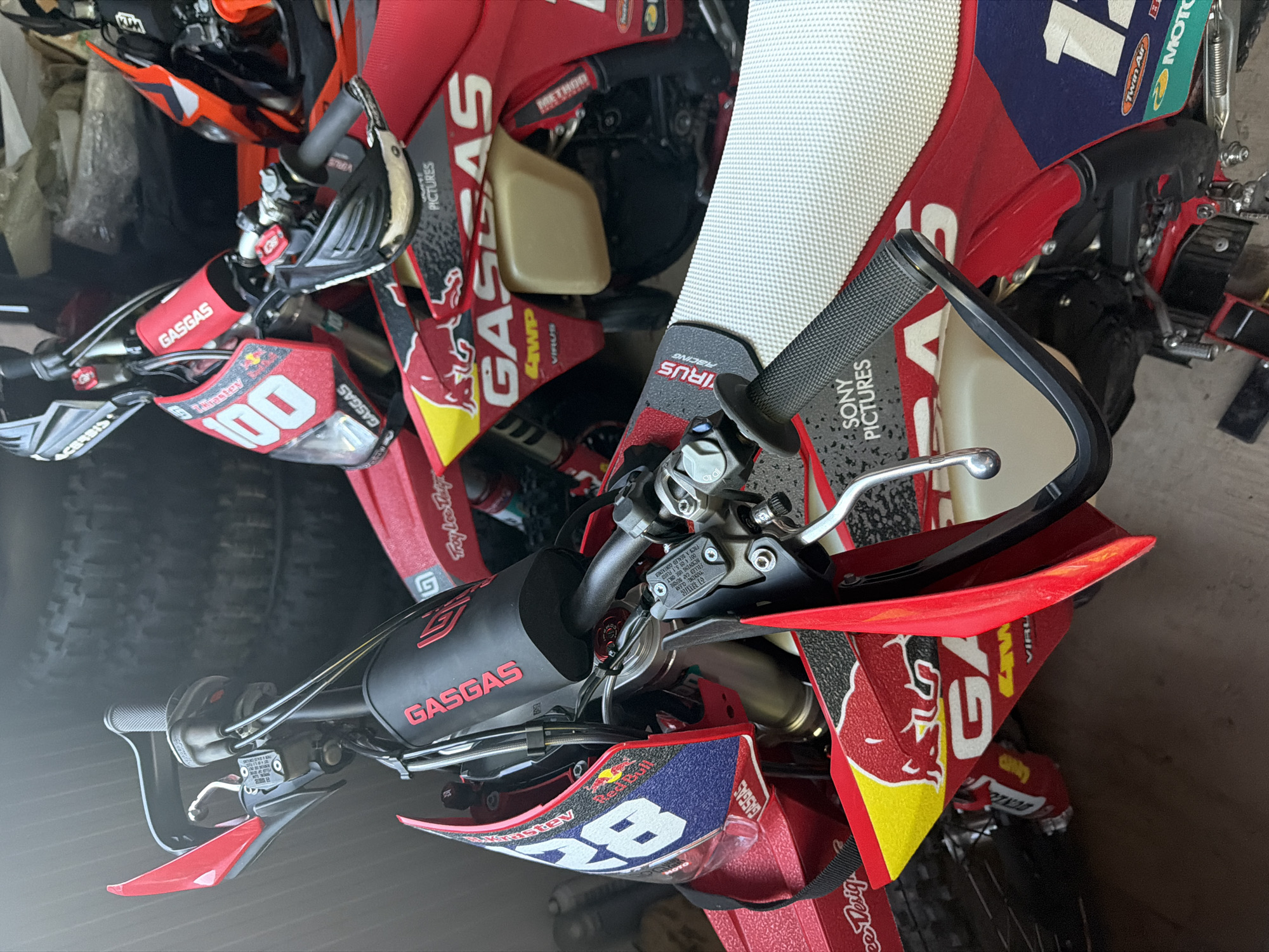 Two red and white dirt bikes are parked indoors, possibly in a garage or storage unit, with stacks of tires in the background. The bike in the foreground has "GASGAS" branding visible on its handlebars and frame, along with "Red Bull" graphics and the number "28" on its front plate. The bike in the background also features "GASGAS" branding and the number "100" on its front plate. The scene appears to be staged, as the bikes are clean and well-maintained. There are no people present in the image, and no discernible actions are taking place. The environment is dimly lit, suggesting an indoor setting with artificial lighting. The location is not specified in the image, and therefore, it cannot be determined if it is in Kazanlak, Bulgaria.