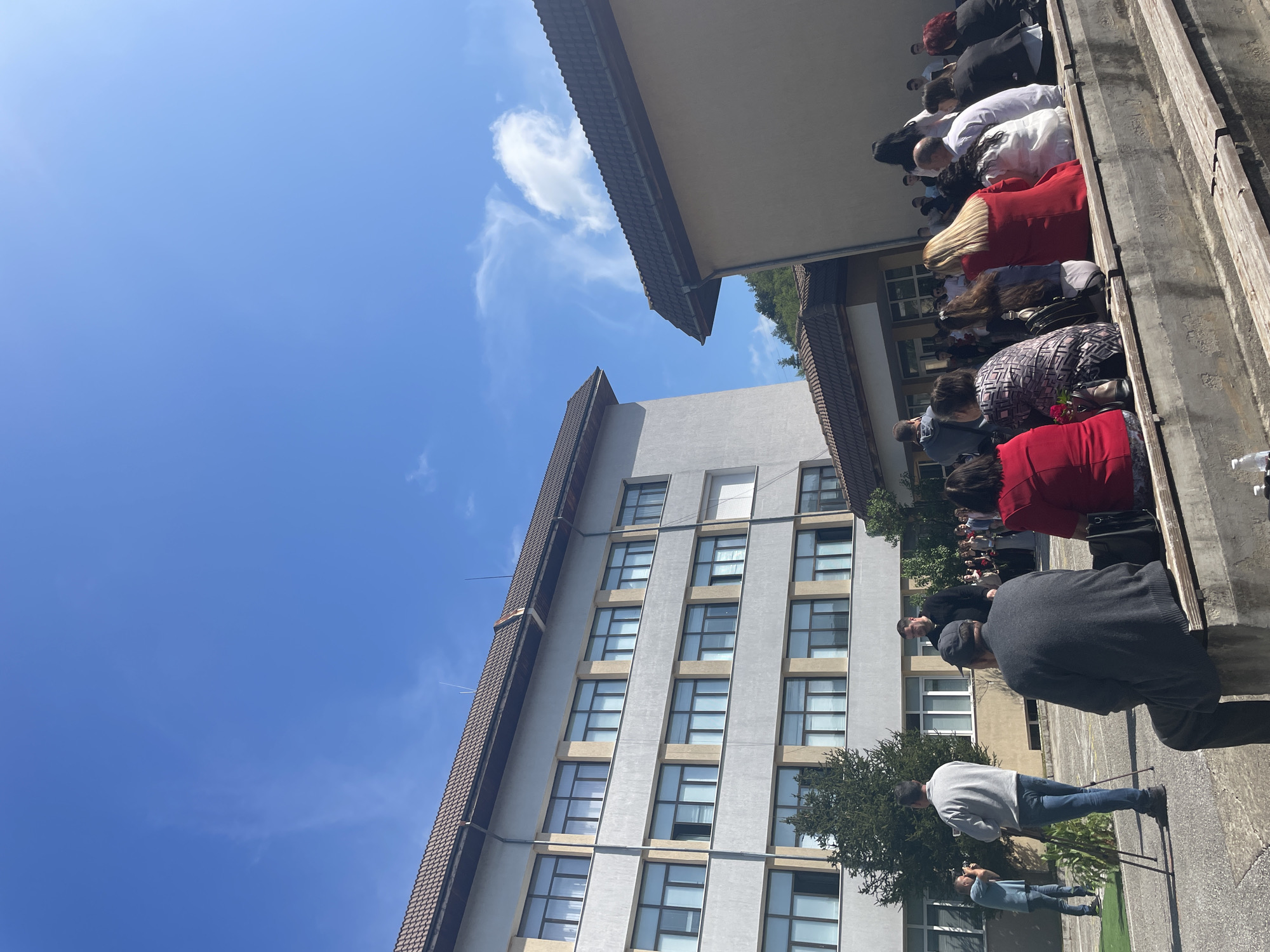 A group of people are standing in front of a building under a clear, blue sky in Zlatograd, Bulgaria. The building is tall and gray with many windows. The people are wearing various clothing, including red, white, black, and gray colors. They appear to be bowing their heads. Some individuals are taking photos. The scene includes architectural elements and trees.