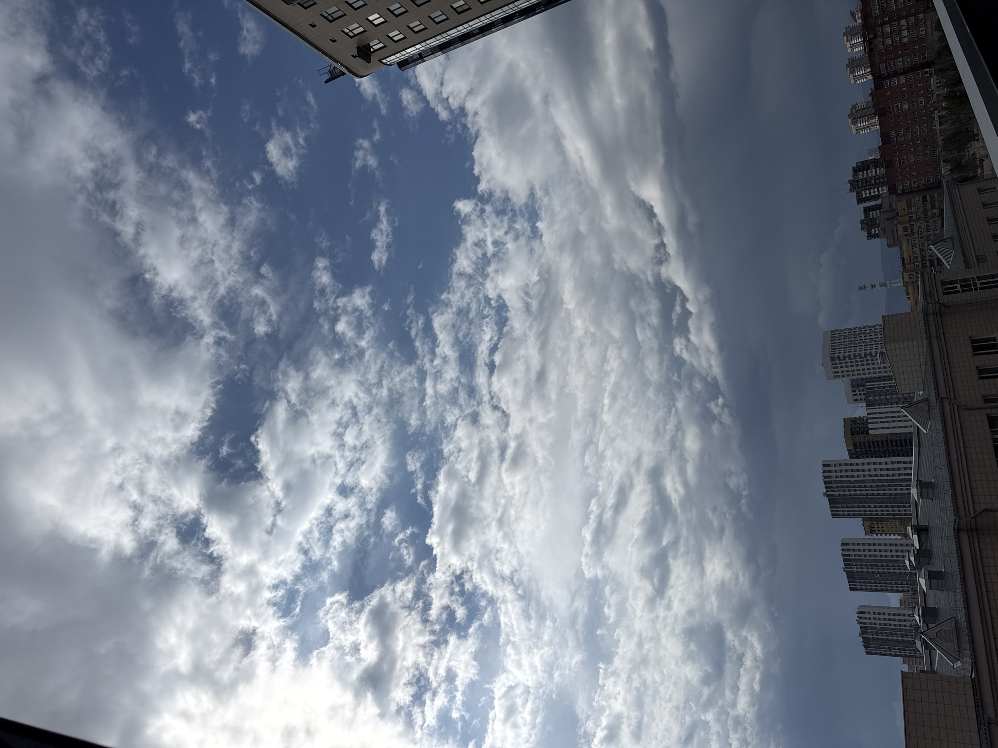 The image captures a sky filled with scattered cumulus clouds, ranging from bright white to shadowed gray, indicating varying cloud thickness and sunlight penetration. Through the gaps in the clouds, patches of blue sky are visible. Along the edges of the frame, tall buildings are situated, suggesting an urban environment. These buildings vary in architectural style, with some appearing to be modern high-rises and others exhibiting older, more traditional designs. The picture orientation is rotated counterclockwise, possibly taken looking upward from street level or an elevated position between buildings. It might be located in Krasnoyarsk, Russia. The specific details of the surrounding urban environment require additional context or a broader perspective within the image to ascertain more granular information.