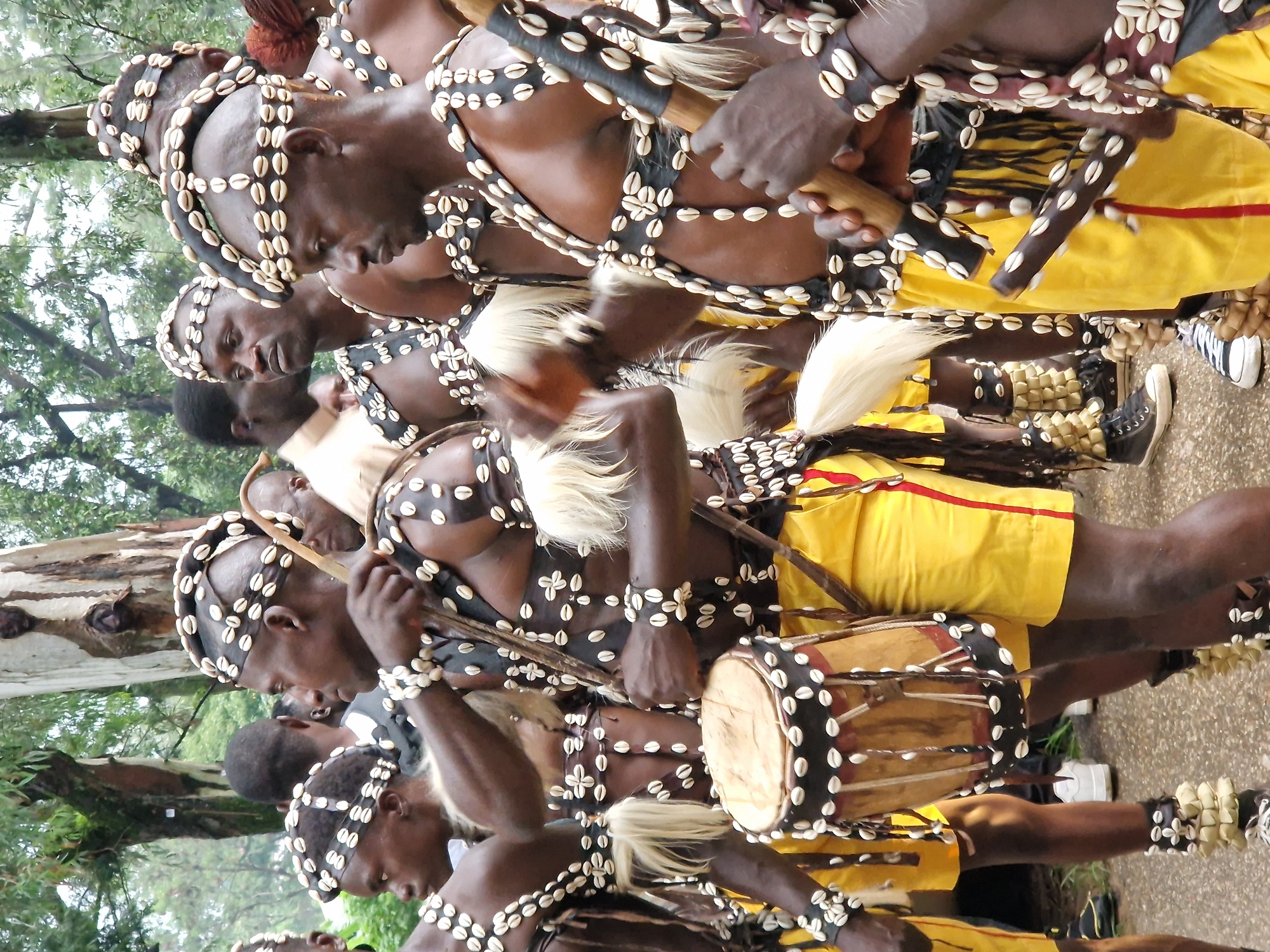 This image depicts a group of men in traditional attire participating in an outdoor event. The men are adorned with elaborate headdresses and body ornamentation made of shells and dark fabric. They are also wearing yellow shorts with red trim. Several individuals are holding wooden sticks, and one man is holding a drum. The scene appears to be set in a natural environment with trees and foliage visible in the background, suggesting a location outside of urban density. The ground is paved, consistent with a street or pathway. The overall atmosphere suggests a cultural celebration or performance. Based on the presence of the traditional regalia, it is plausible that this event is taking place in Jos, Nigeria, a city known for its diverse cultural heritage and vibrant traditional practices.
