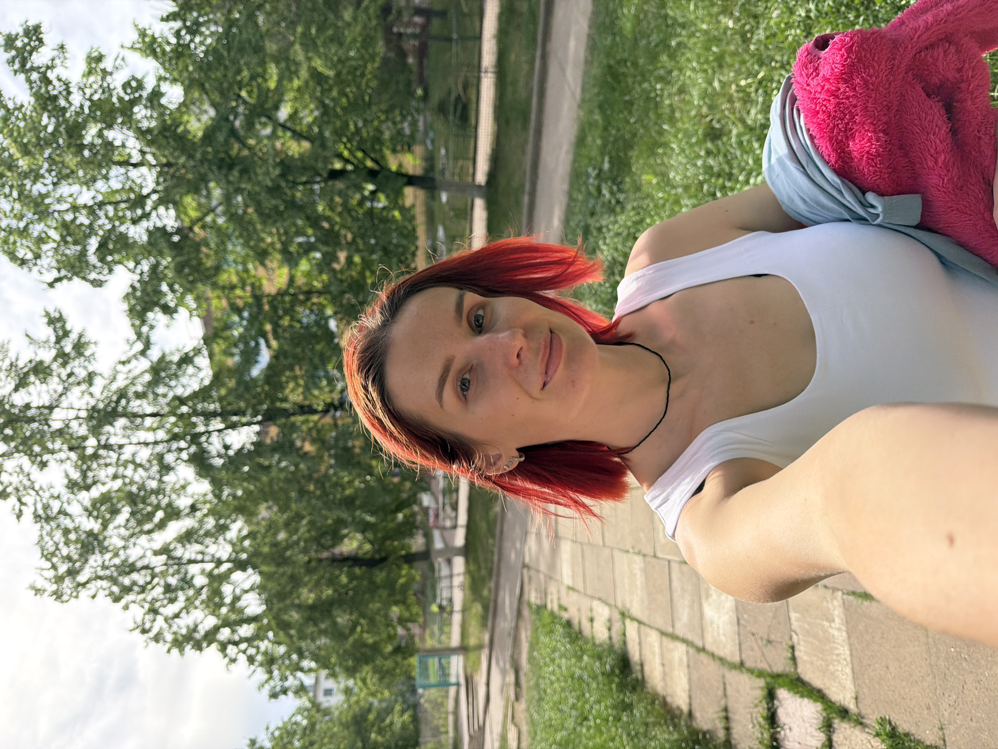 A woman with red hair wearing a white tank top is taking a selfie outdoors. The location appears to be a park or green space in the city of Chaikovsky, Russia, evidenced by the presence of trees, grass, and a paved walkway. The woman has a pink fuzzy item and a light blue garment draped over her shoulder.